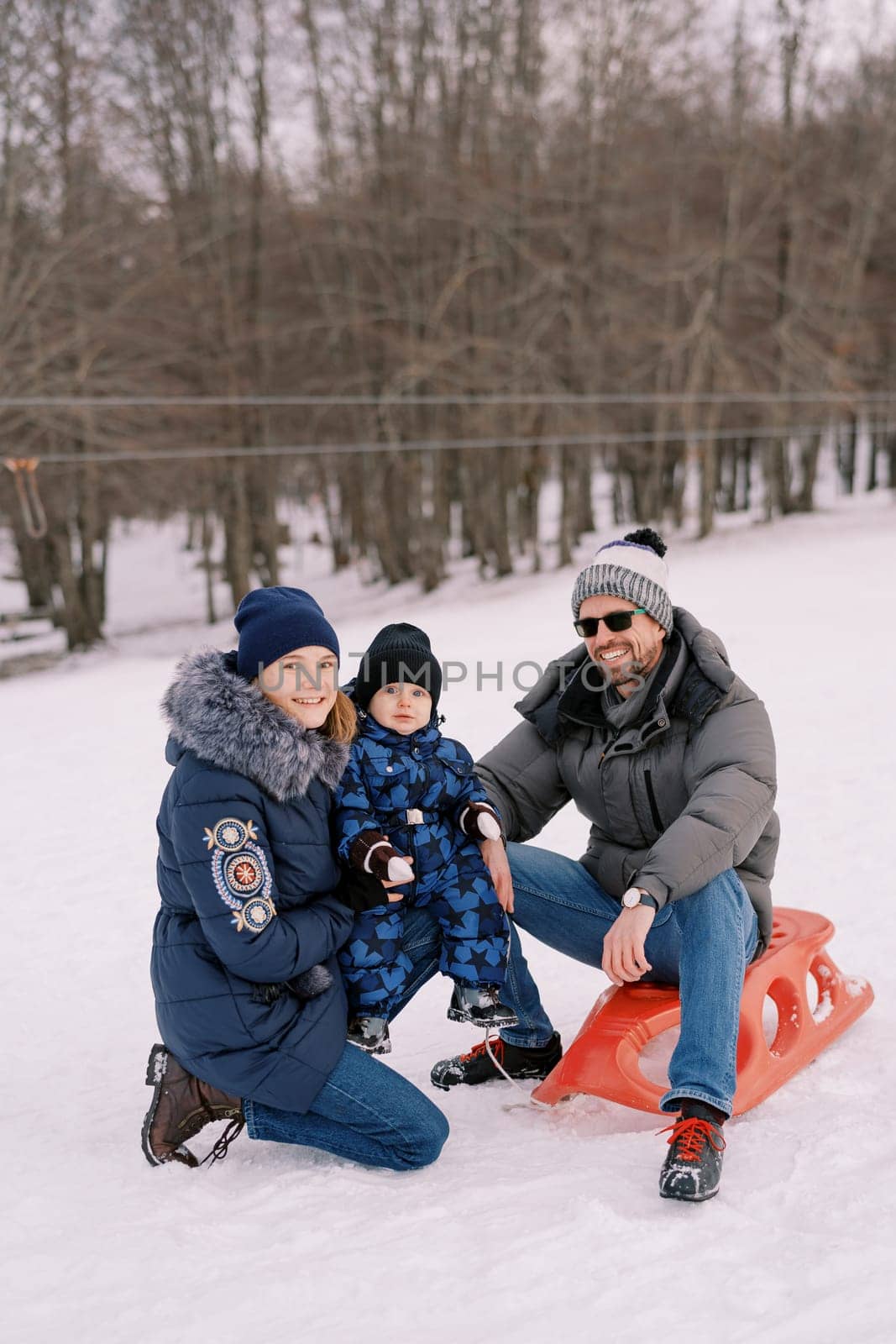 Smiling dad sitting on sled next to squatting mom with little boy on lap. High quality photo