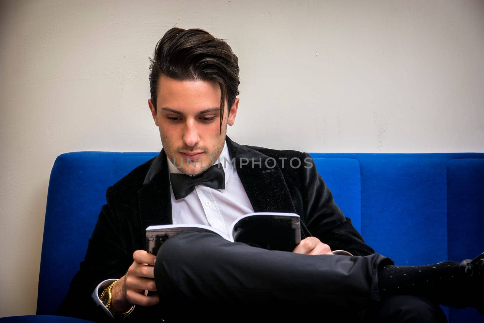 A man in a tuxedo sitting on a blue couch reading a book