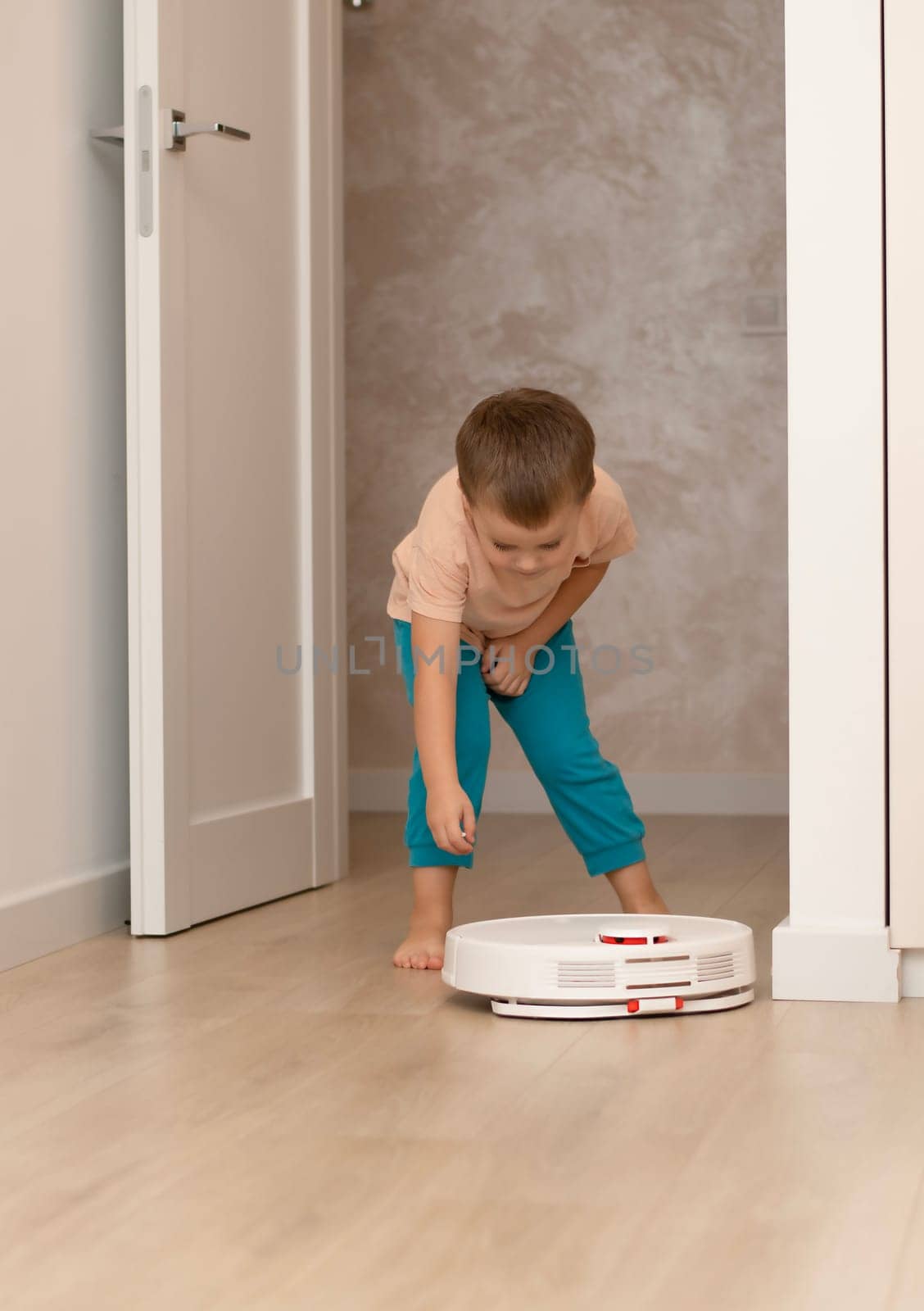 Cleaning concept. A little funny Caucasian boy, 4 years old, throws torn paper in small pieces onto the floor for cleaning and suction by a white robot vacuum cleaner. Plays happily in the home interior.