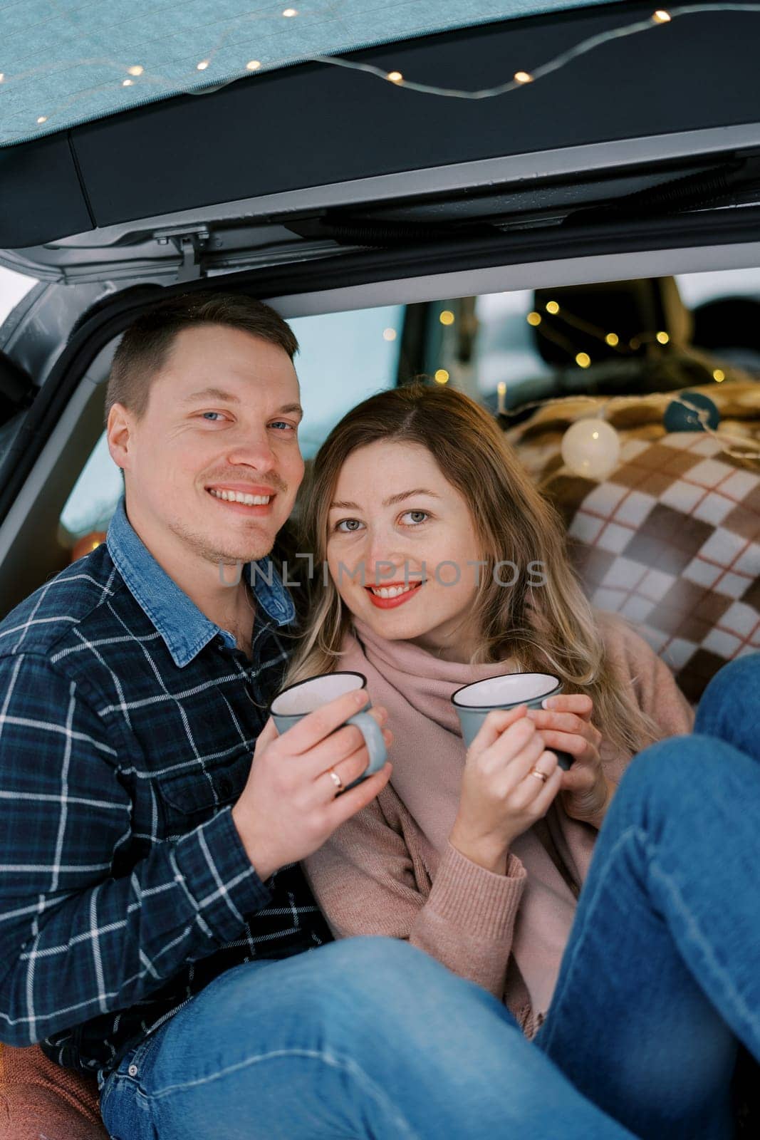 Smiling woman leaning against man sitting with mugs of coffee in car trunk. High quality photo