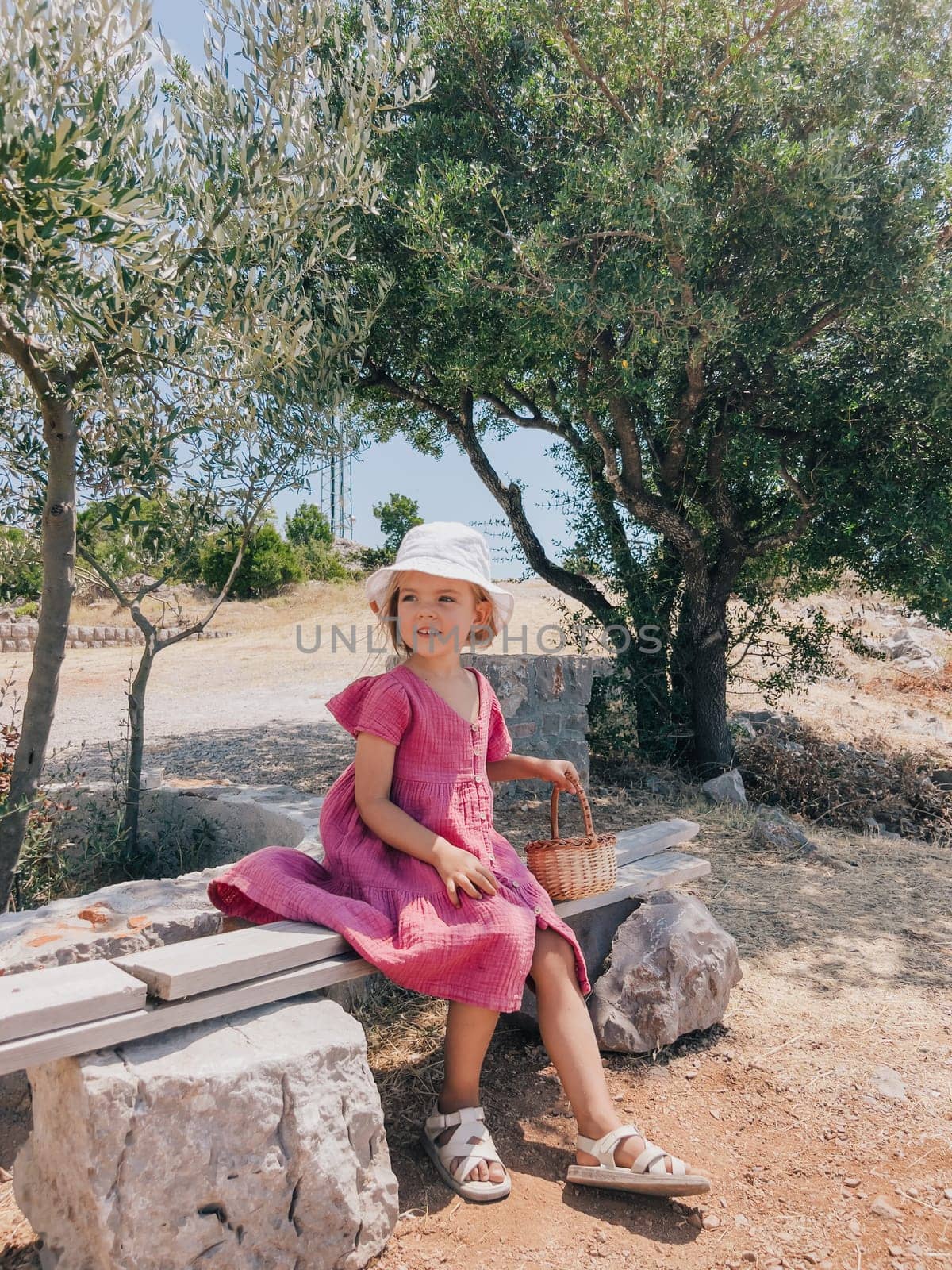 Little smiling girl with a basket sits on a bench near a tree. High quality photo