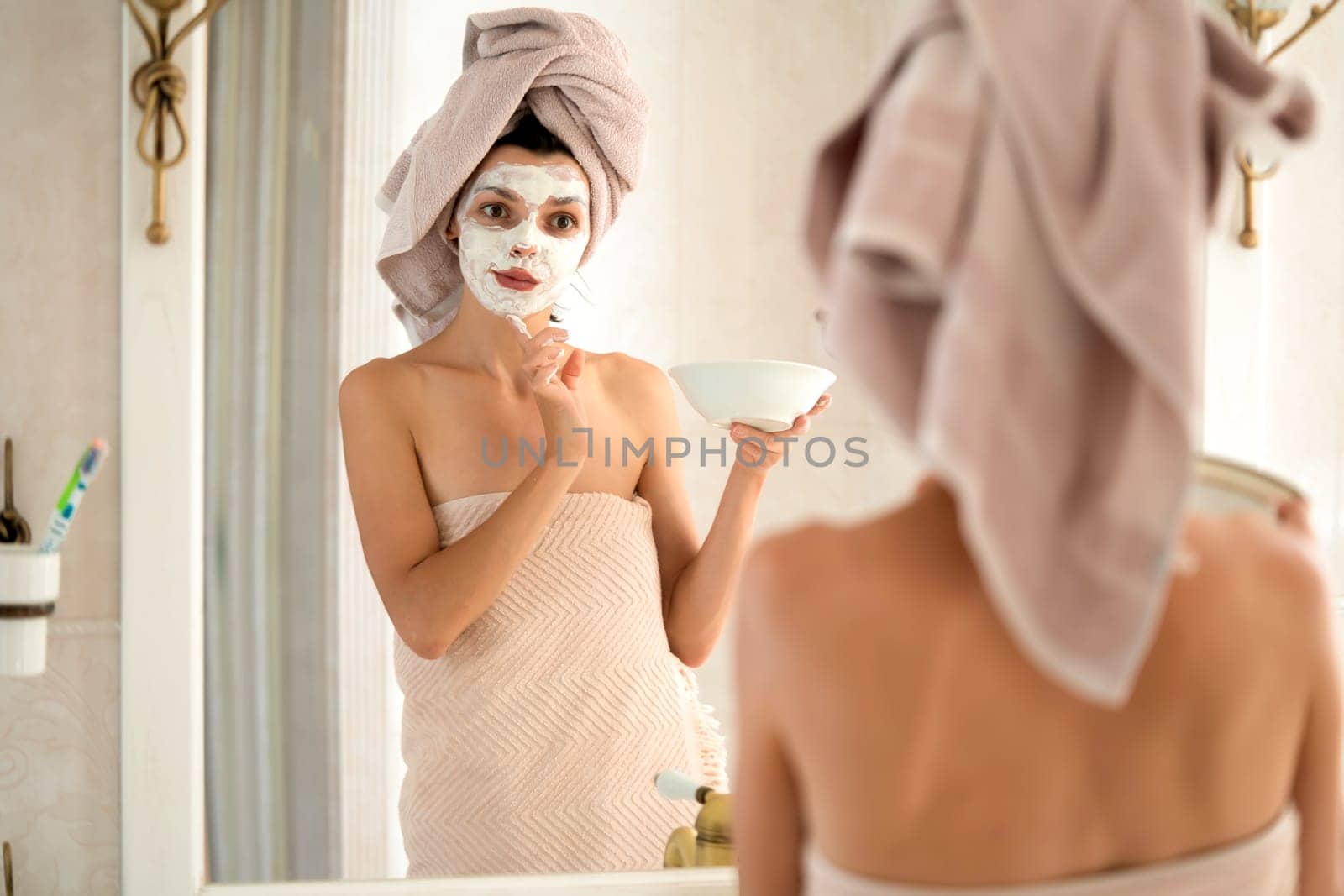 A young girl in a towel on her head and body stands near the mirror in the bathroom and applies a clay mask to her face, the woman takes care of her health and beauty.