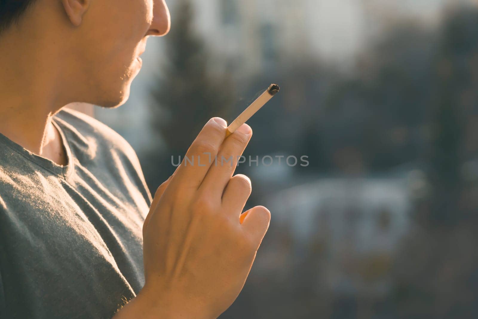 A young man smokes, holds a cigarette in his hand and inhales the smoke while standing at the window early in the morning in the sun, close-up view.