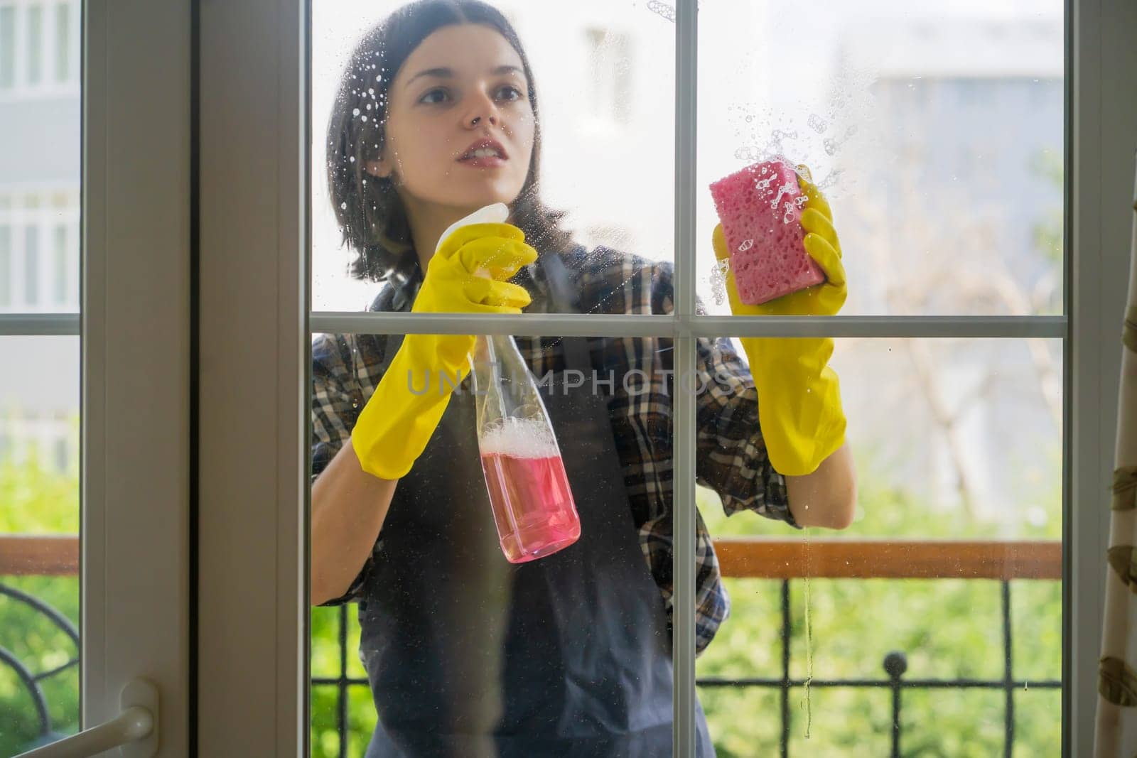 A young girl in yellow rubber gloves and a uniform washes the windows thoroughly with a detergent, wiping them dry with a rag, the woman is cleaning the house.