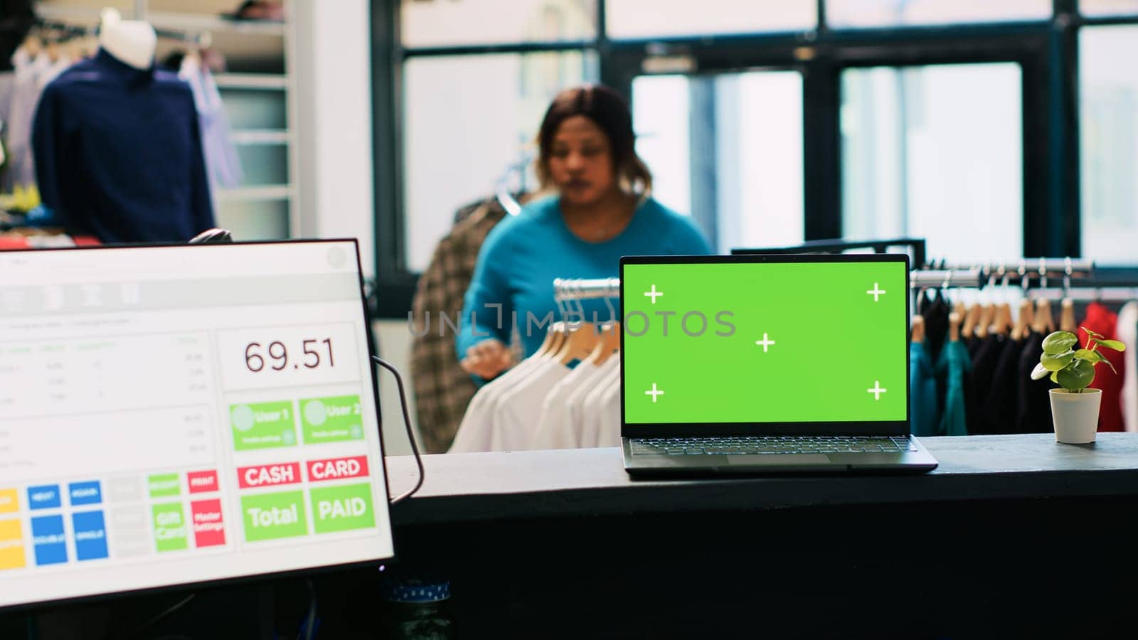 Laptop computer with chroma key green screen mock up display standing on counter desk in clothing store. In background diverse customers shopping for fashionable merchandise in modern boutique