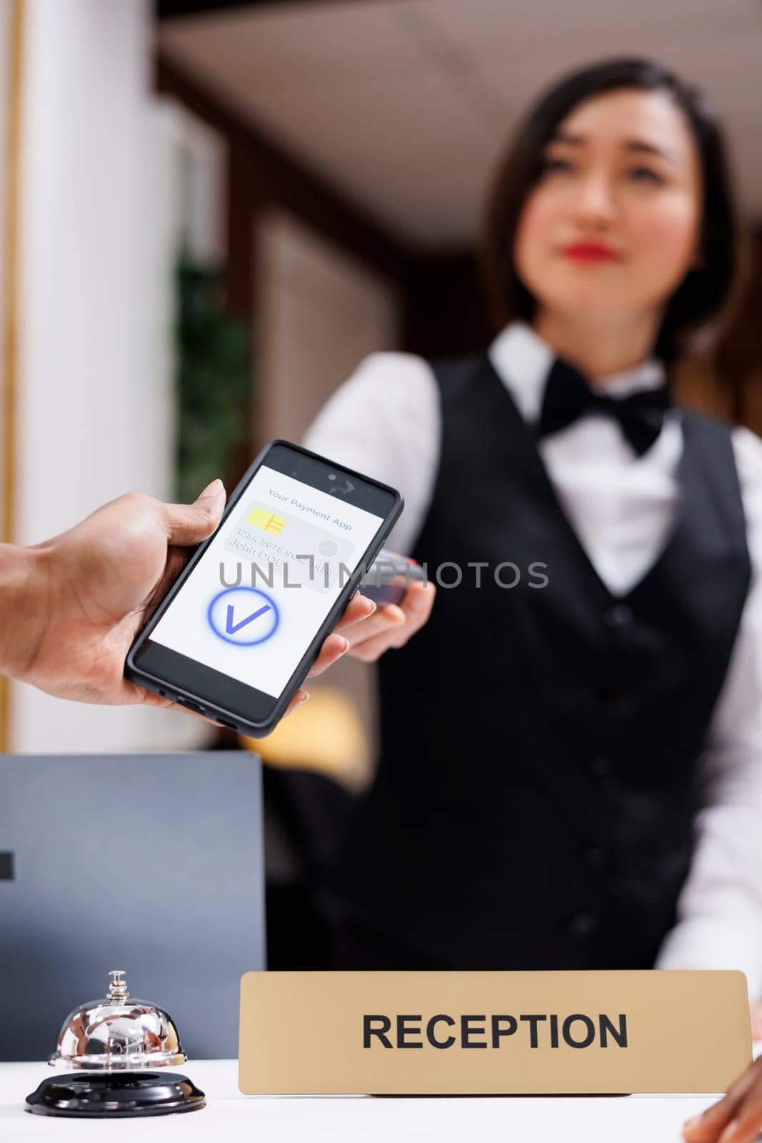 Man paying with mobile phone and nfc at pos terminal, receptionist helping people with check in process. Asian front desk staff welcoming guests, adult making payment for room. Close up.