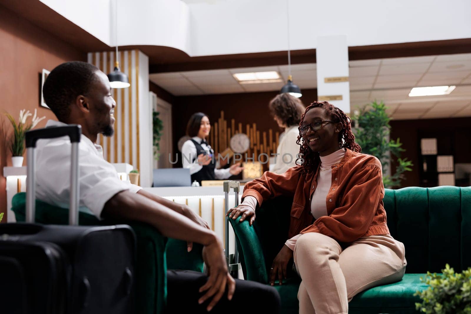 Young couple waiting to do check in at hotel reception, relaxing on couch in luxury lounge area. African american people sitting in modern resort lobby before filling in registration forms.