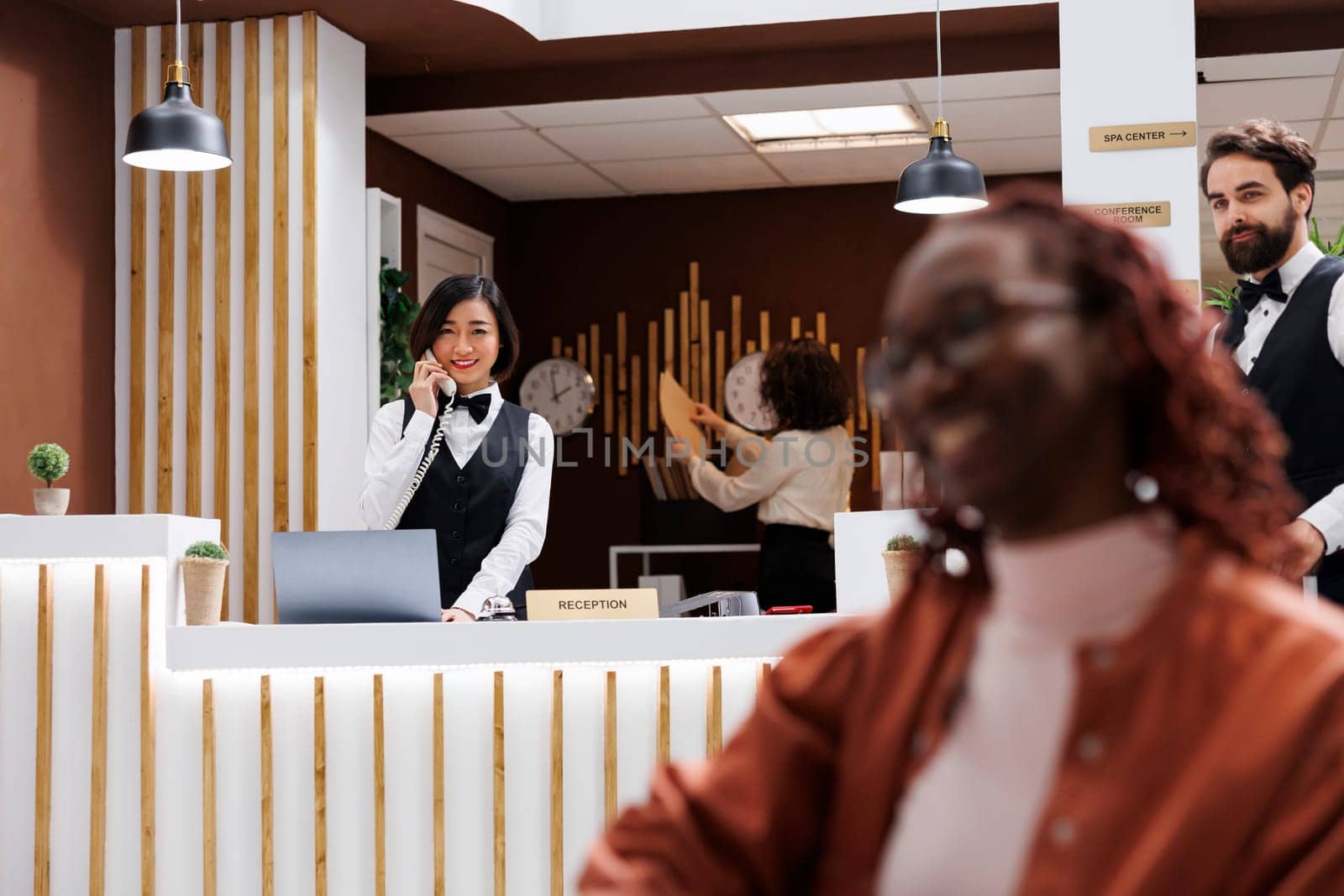 Employee making online room booking on phone, talking on landline call to note room reservation in hotel records. Female reception worker helping future resort guests with accommodation.