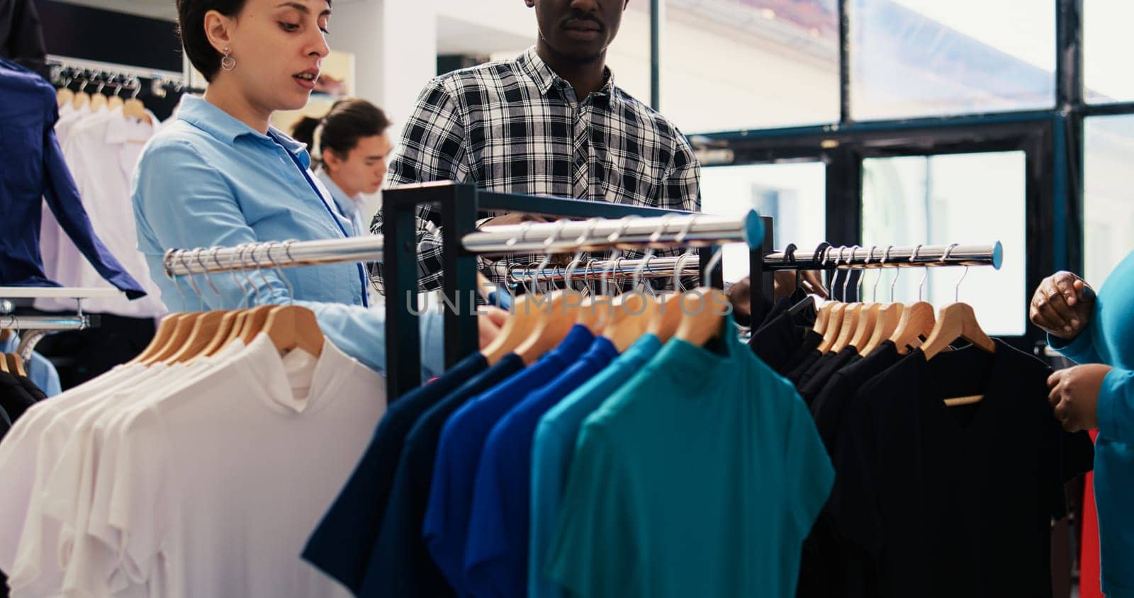 Stylish employee showing black shirt to couple, checking new fashion collection in modern boutique. African american customers shopping for trendy merchandise and accessories in clothing store