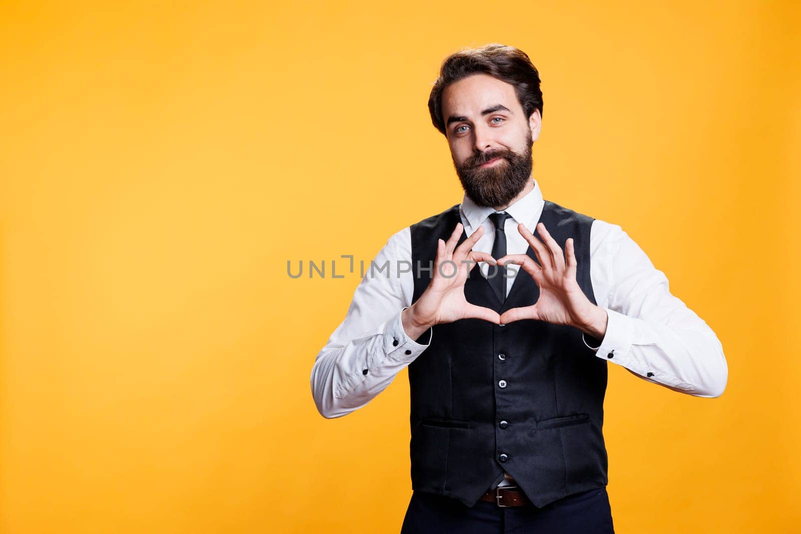 Butler presents heart shaped sign in studio, working at restaurant and showing true emotions on camera. Young adult acting romantic and expressing love with symbol, sincere feelings.