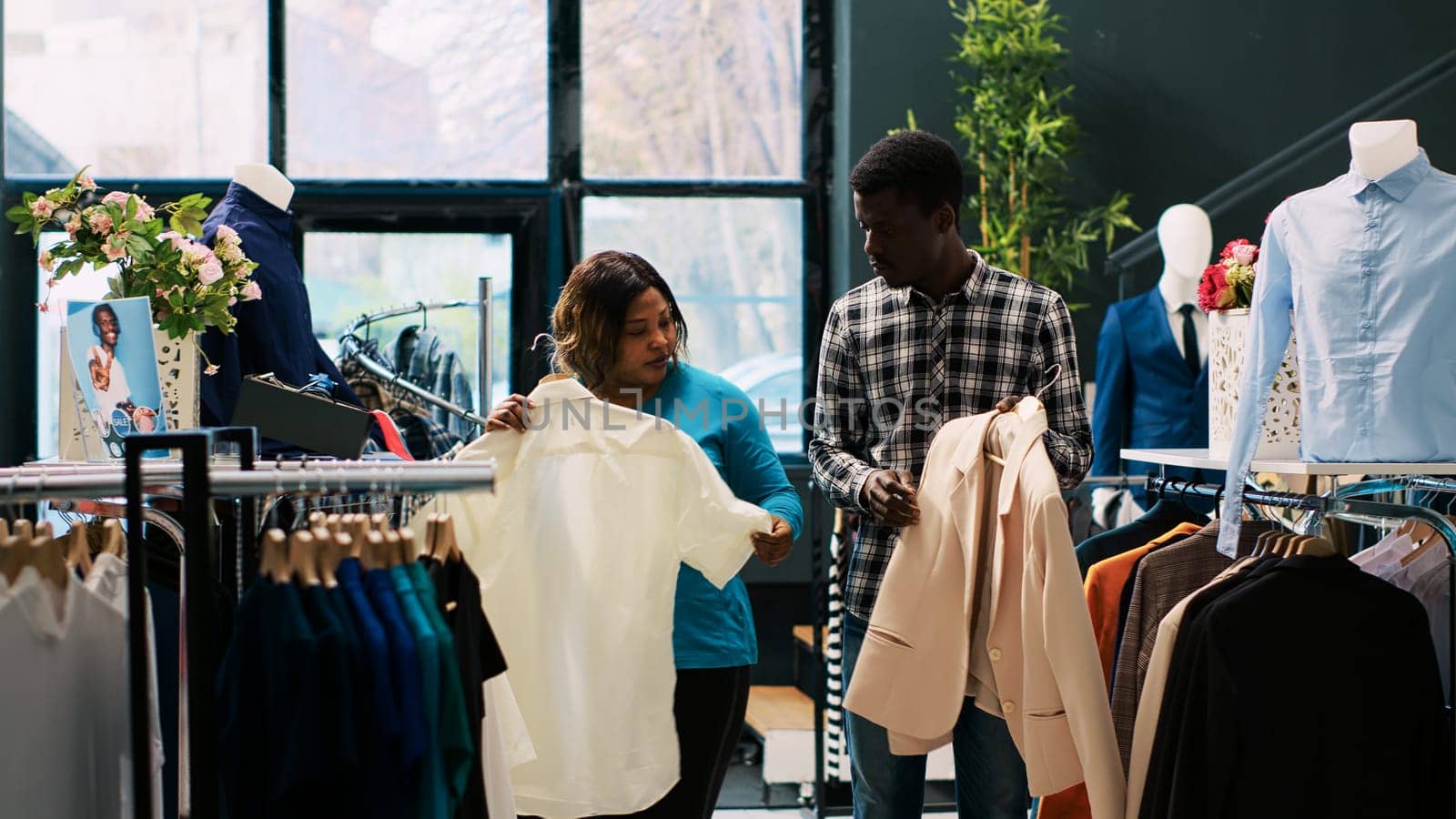 African american couple looking at trendy clothes, shopping for casual wear in modern boutique. Cheerful customers checking outfit material, buying stylish merchandise for new wardrobe. Fashion concept