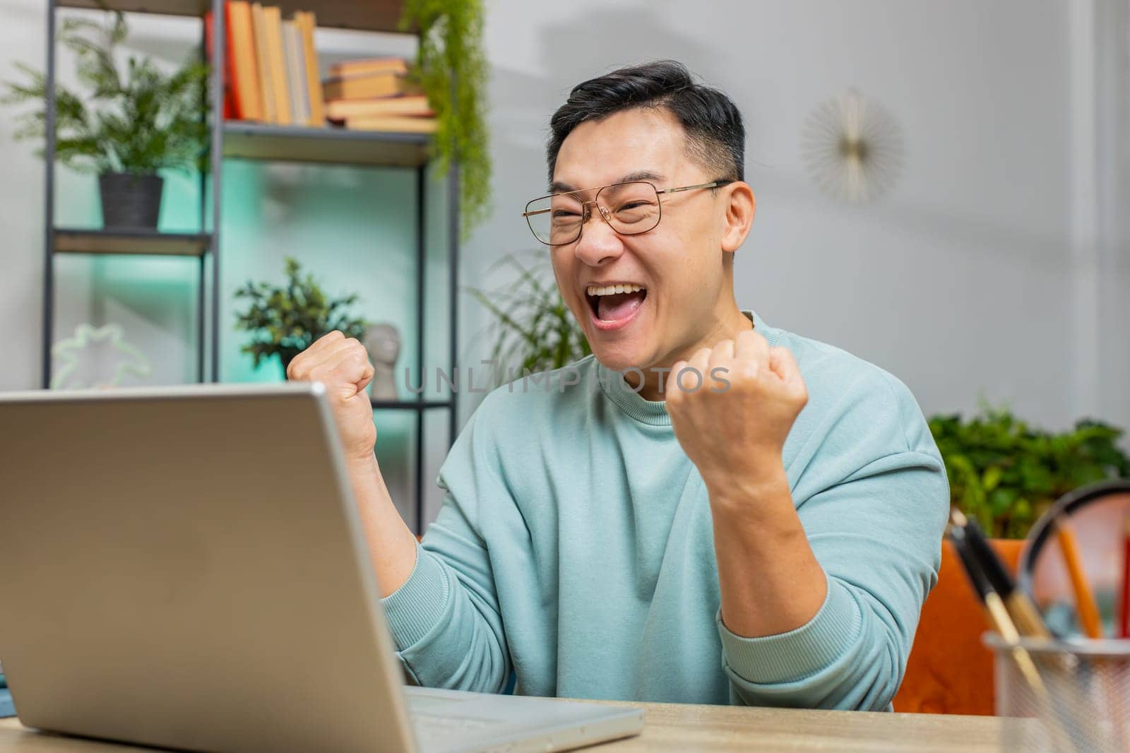 Happy Asian middle-aged mature man using laptop computer, receive mail good news message shocked by sudden victory celebrate lottery jackpot win purchases online shopping play game at home apartment