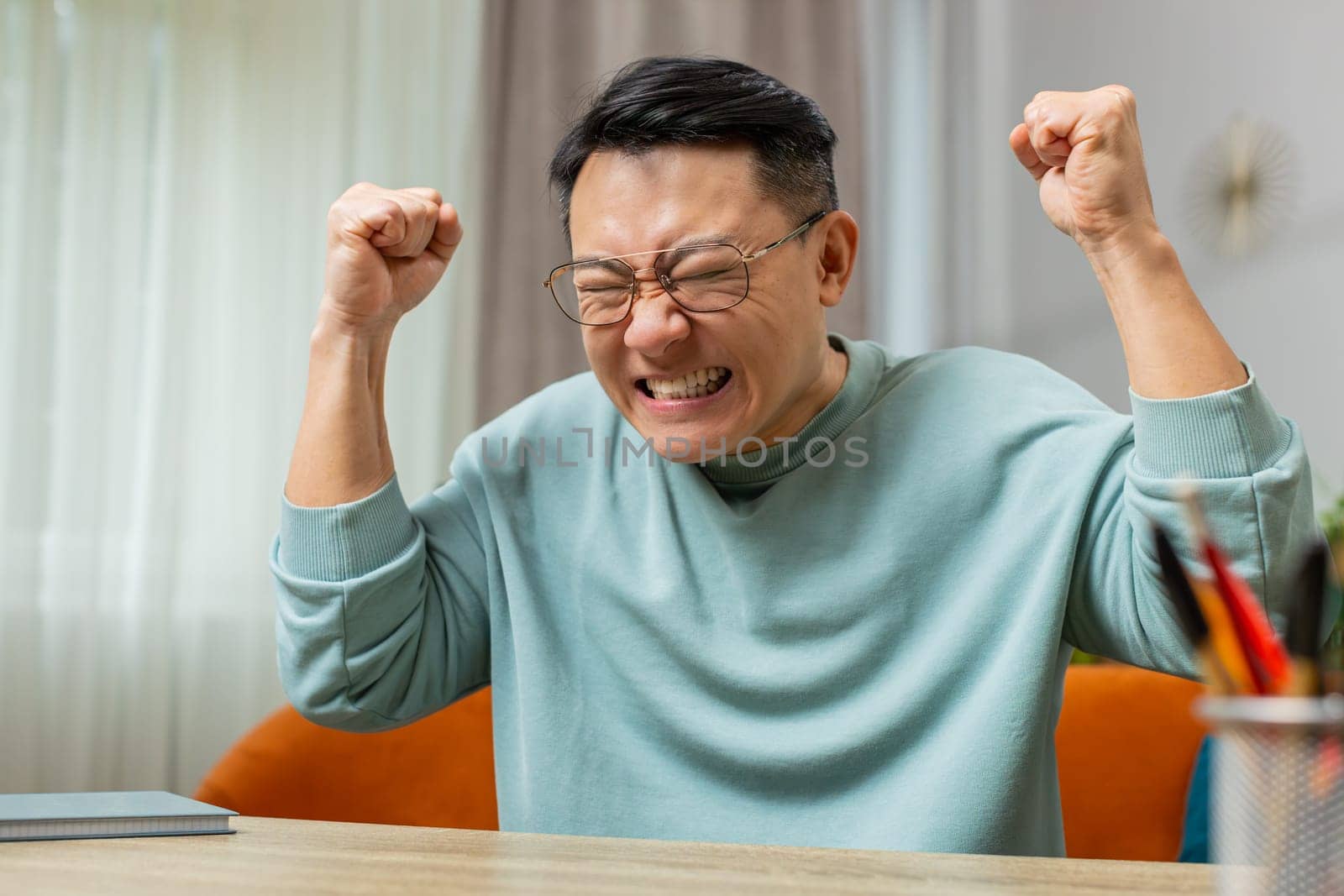 Aggressive Asian mature man screams from stress tension problems panic horror hopelessness fear surprise shock expresses gestures rage nervous. Furious guy at home apartment room sit on couch at table