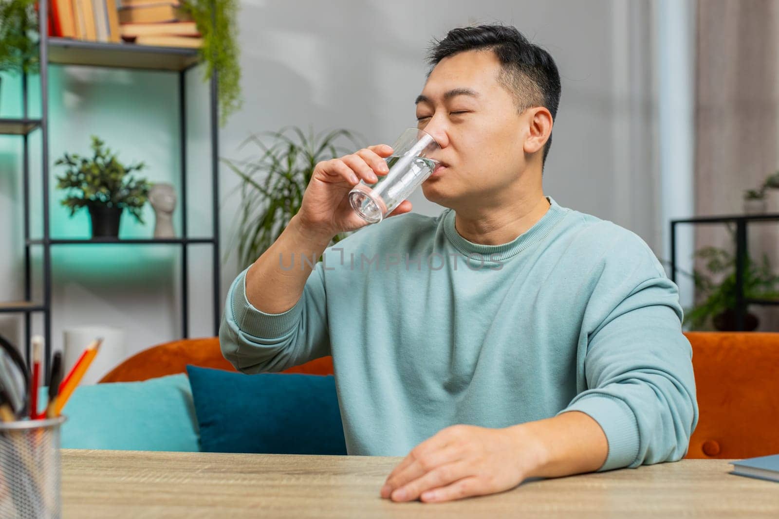 Thirsty Asian man hold glass of natural filtered aqua make sips drinking still water preventing dehydration at home. Water after reverse osmosis filter. Guy with good life habits, healthy, weight loss