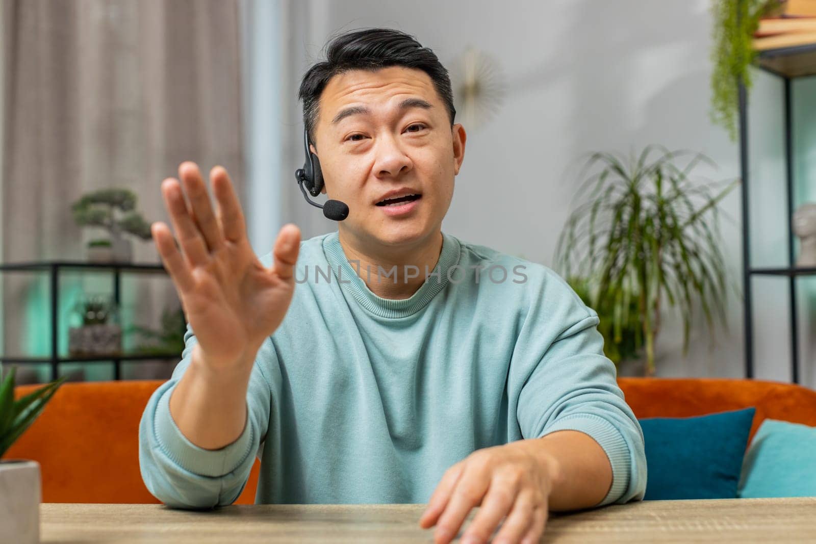 Asian Chinese man wearing headset, freelance worker call center or support service operator helpline. Chinese guy having talk with client or colleague communication support waving hello alone at home