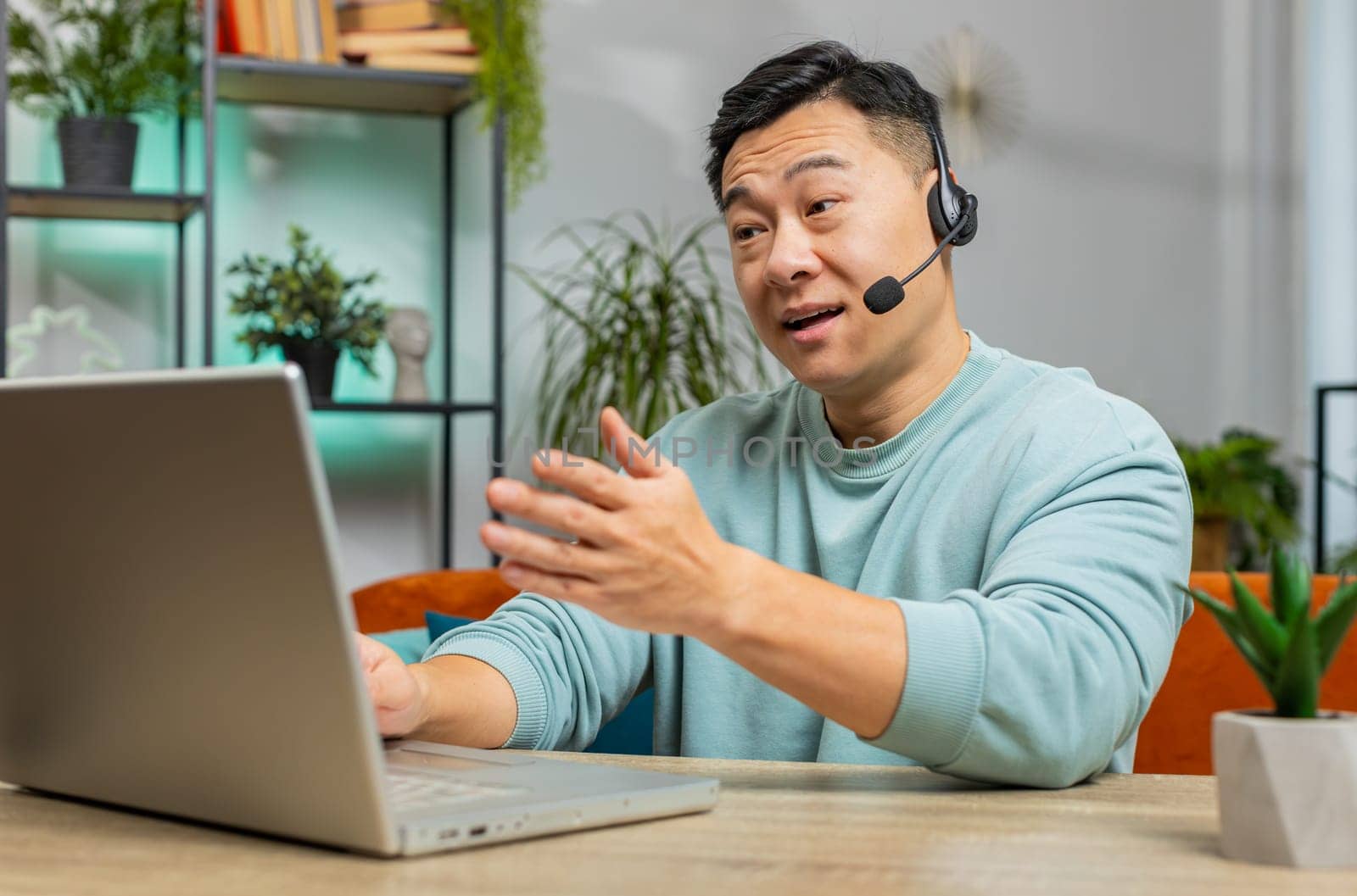 Asian man wearing headset, freelance worker, call center or support service operator helpline, having talk with client or colleague communication support at home room. Guy sits on couch at table desk