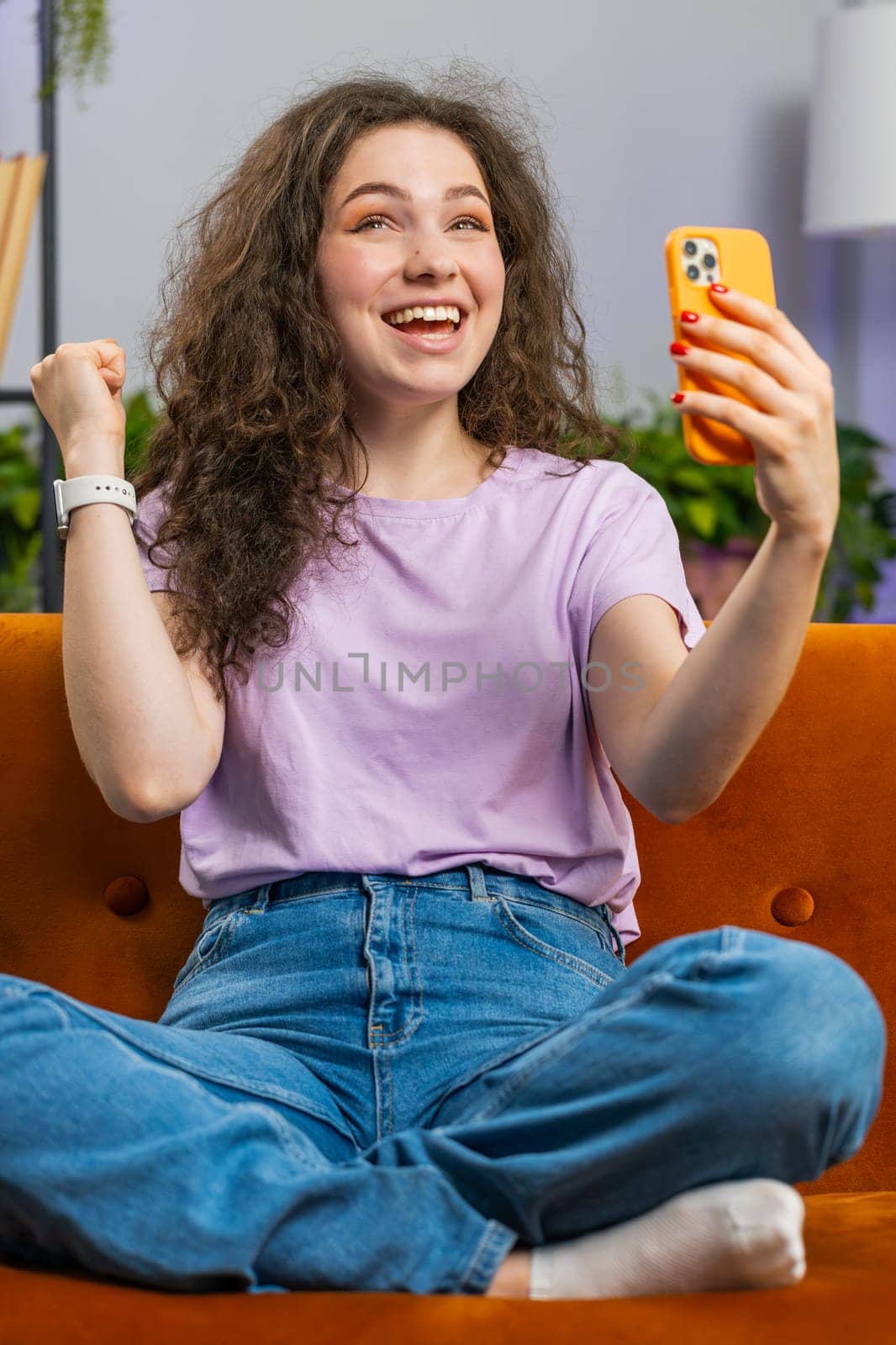 Happy young woman use mobile smartphone typing browsing say Wow yes found out great big win good news celebrate lottery jackpot doing winner gesture. Adult girl at home in room lying on sofa. Vertical