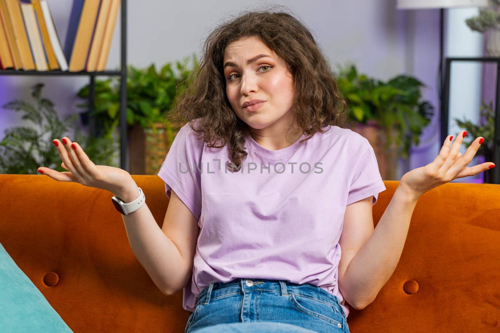 I dont know what to say. Confused pretty woman feeling embarrassed about ambiguous question, having doubts, no idea, being clueless and uncertain at home. Portrait of girl sits on sofa in living room