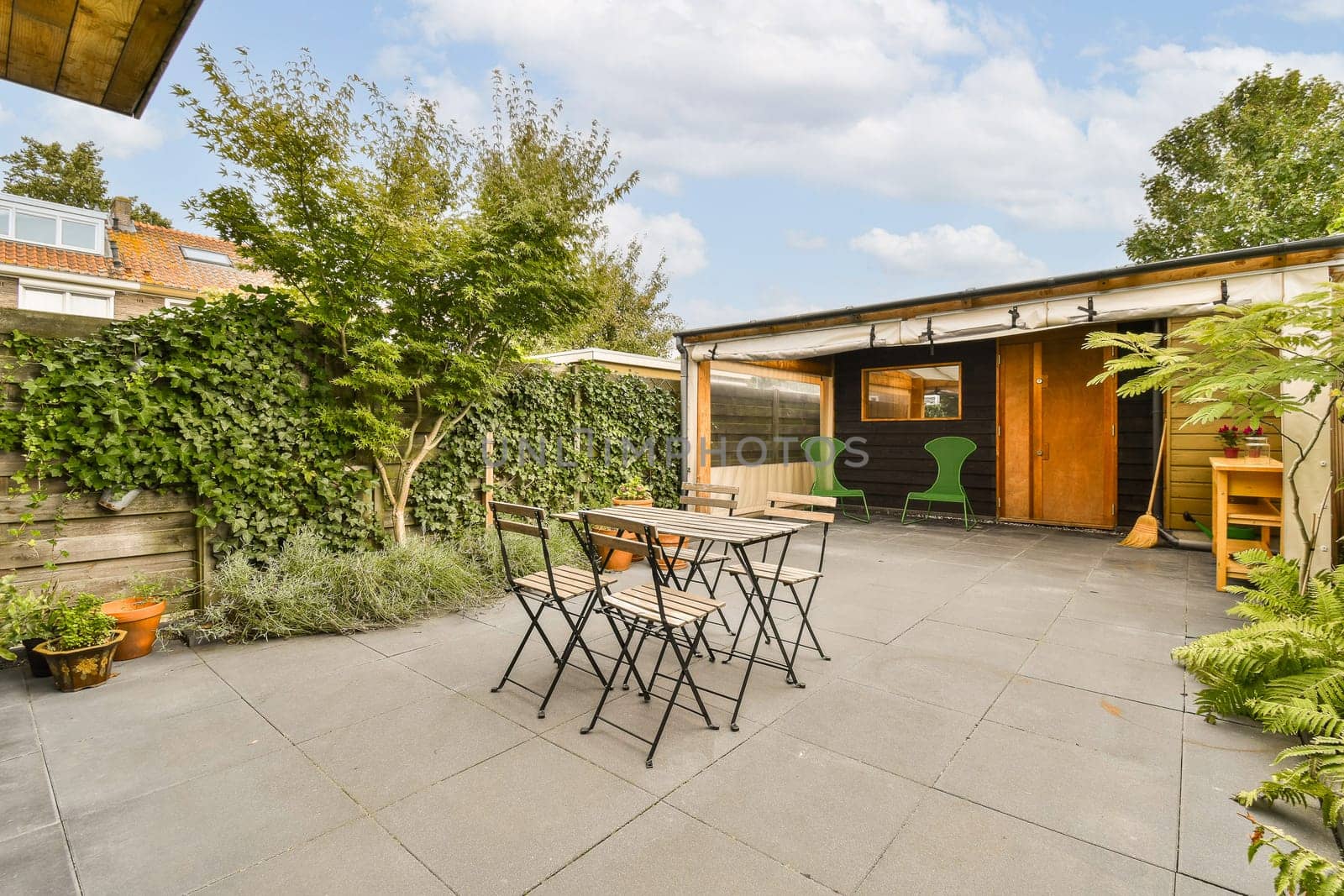 a patio with tables and chairs in the middle of it, surrounded by lush green plants on either side of the house