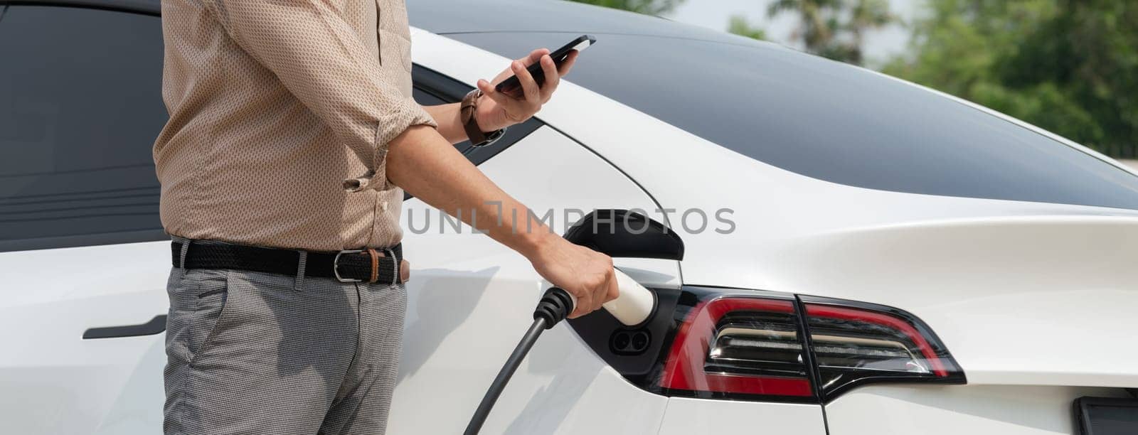 Young man use smartphone to pay for electricity at public EV car charging station green city park. Modern environmental and sustainable urban lifestyle with EV vehicle. Panorama Expedient