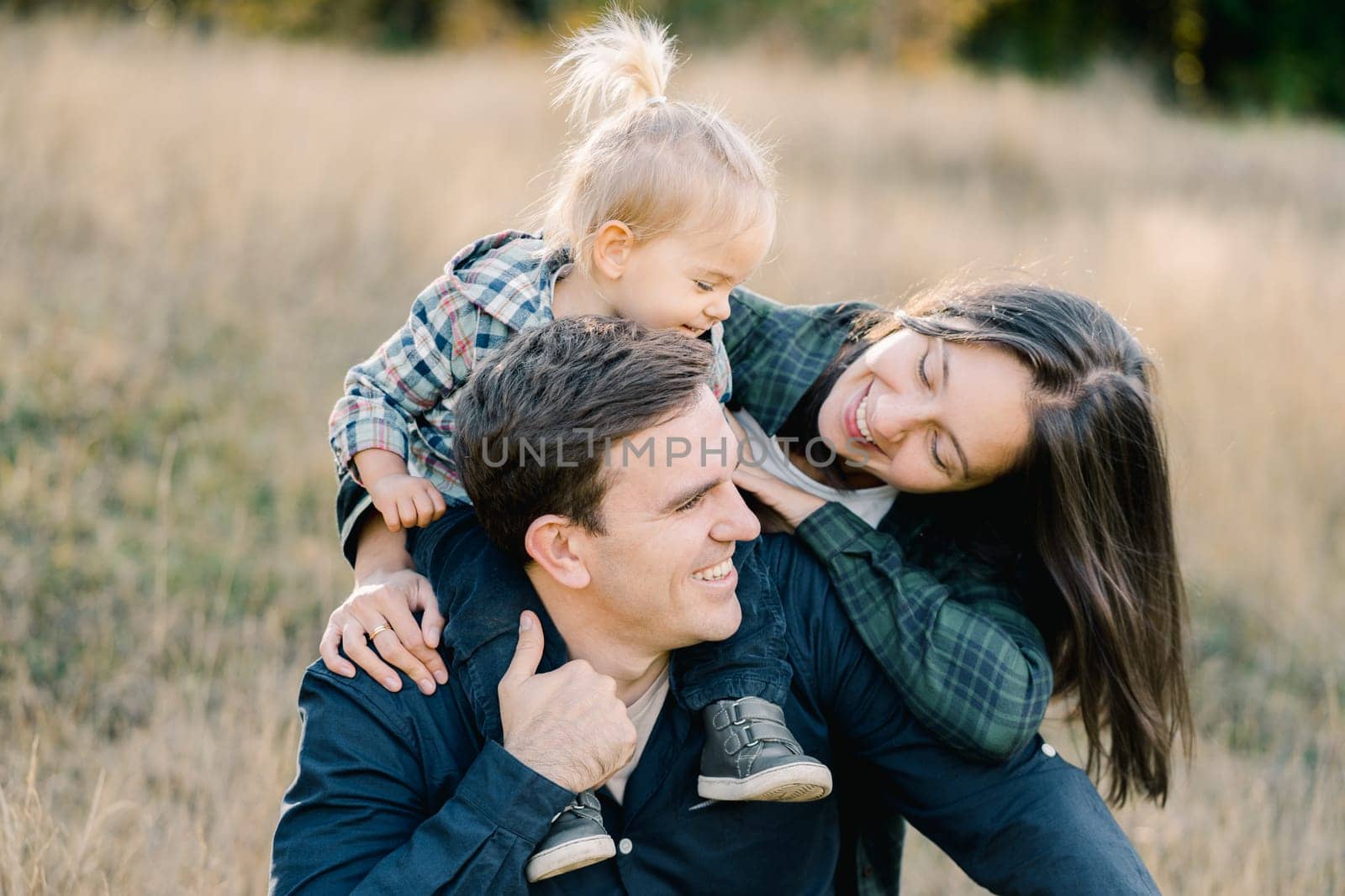 Smiling mom hugging dad with a little girl on his shoulders from behind, looking into his face. High quality photo