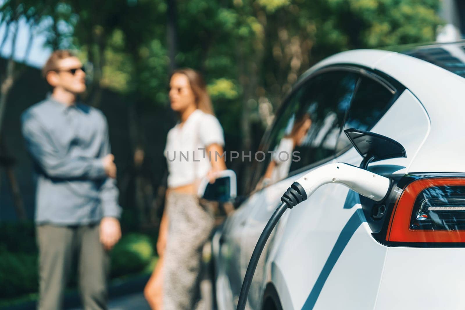 Young couple travel with EV electric car charging in green sustainable city outdoor garden in summer shows urban sustainability lifestyle by green clean rechargeable energy of electric vehicle innards