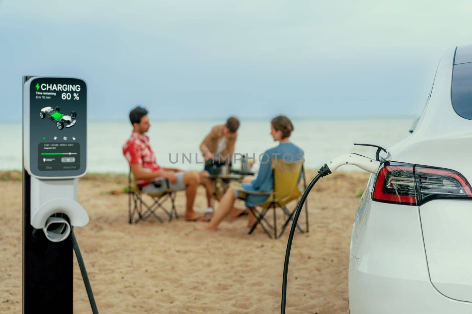 Alternative family vacation trip traveling by the beach with electric car recharging battery from EV charging station with blurred family enjoying the seascape background. Perpetual