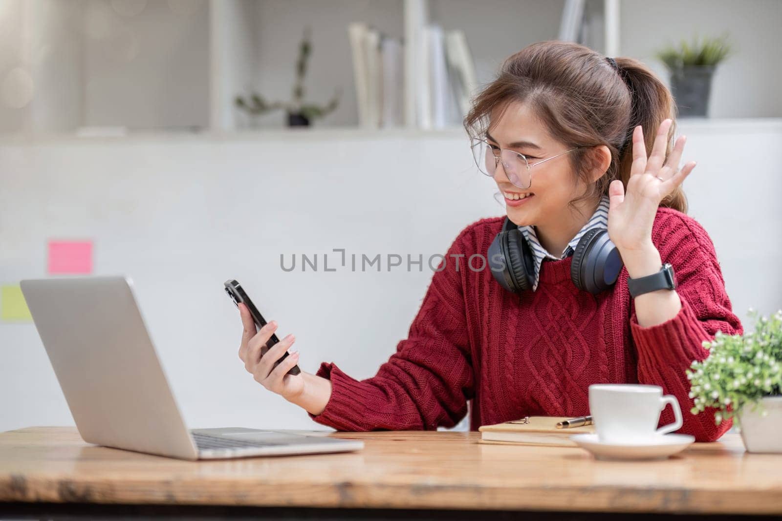 Asian female college student using laptop and phone with headphones while studying. Reading messages and greeting friends via video call.