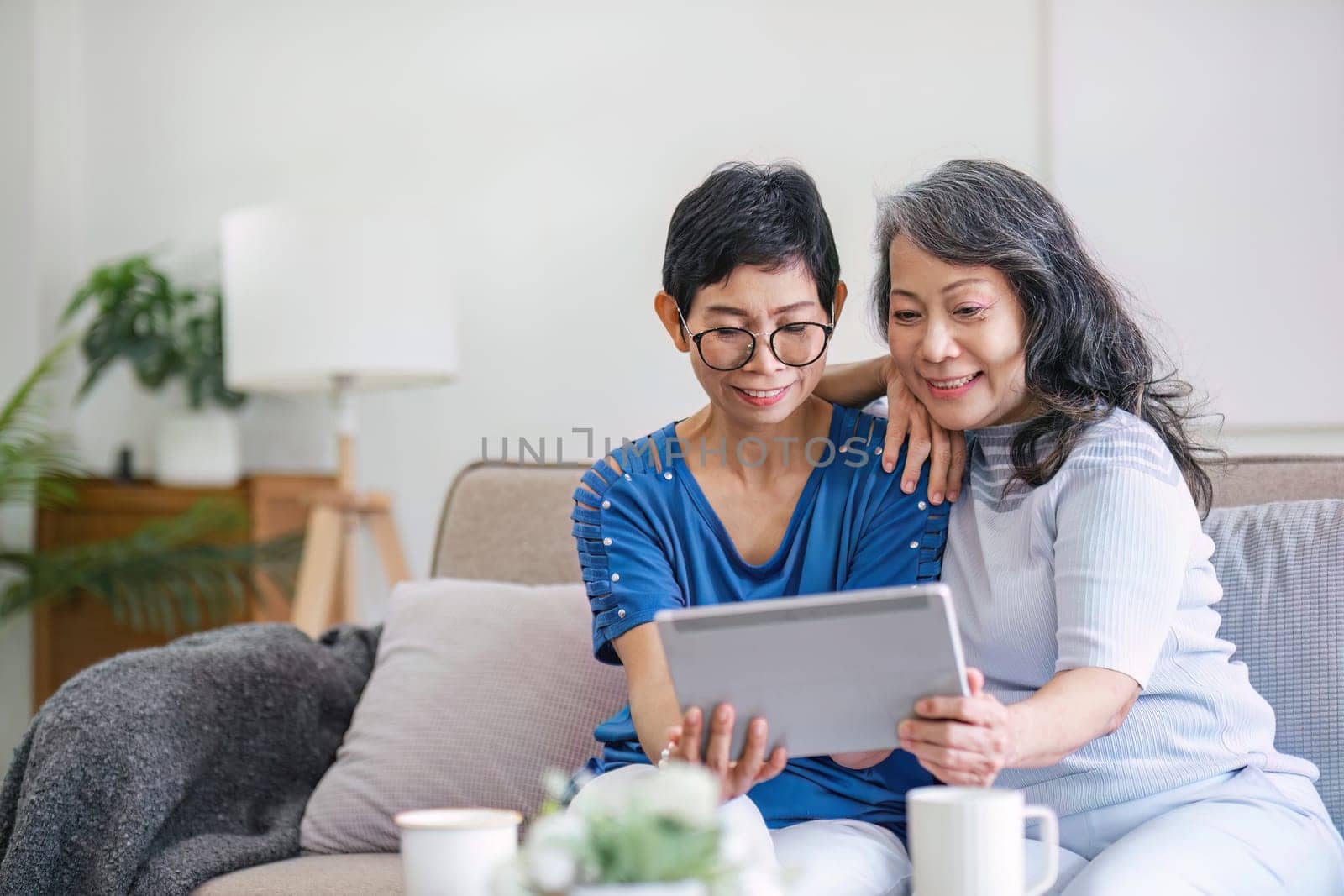 Two old female besties are watching a online media on a tablet while relaxing in the living room together. by wichayada