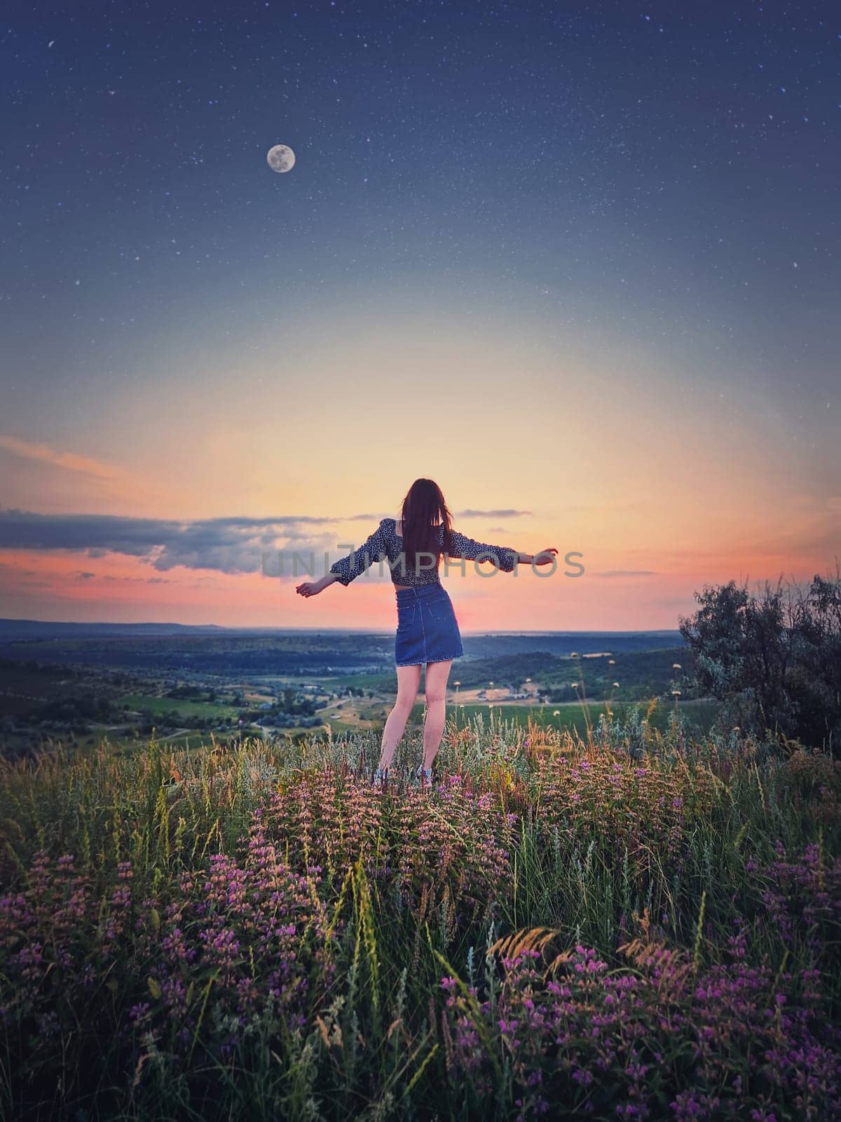 Wild and free youth concept with a relaxed girl, rear view stands with hands outstretched facing the beautiful summer sunset on the top of a hill looking over the valley