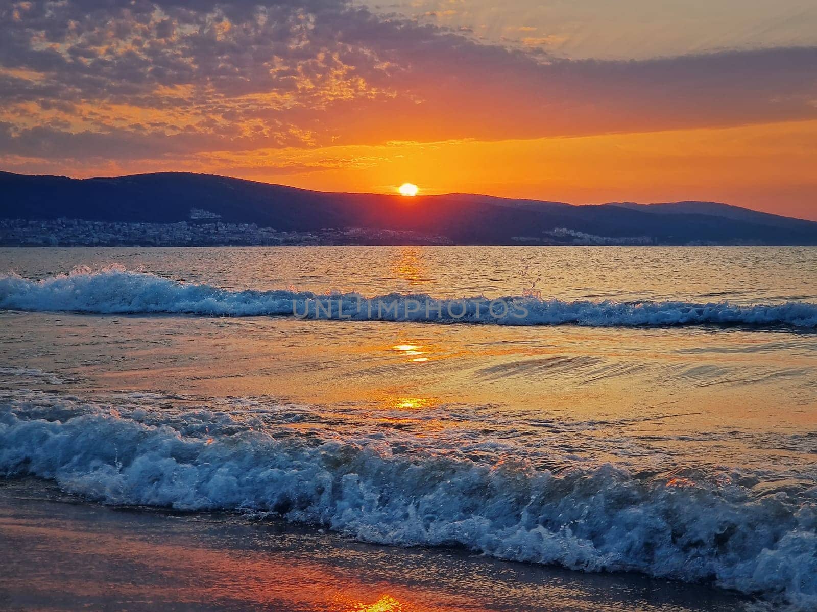Beautiful sunrise at the Bulgarian coastline of Black Sea. Sunny beach resort, dawn scene with the sun rising up the hills on the horizon and foamy waves washing the sand shore. Summer vacation view
