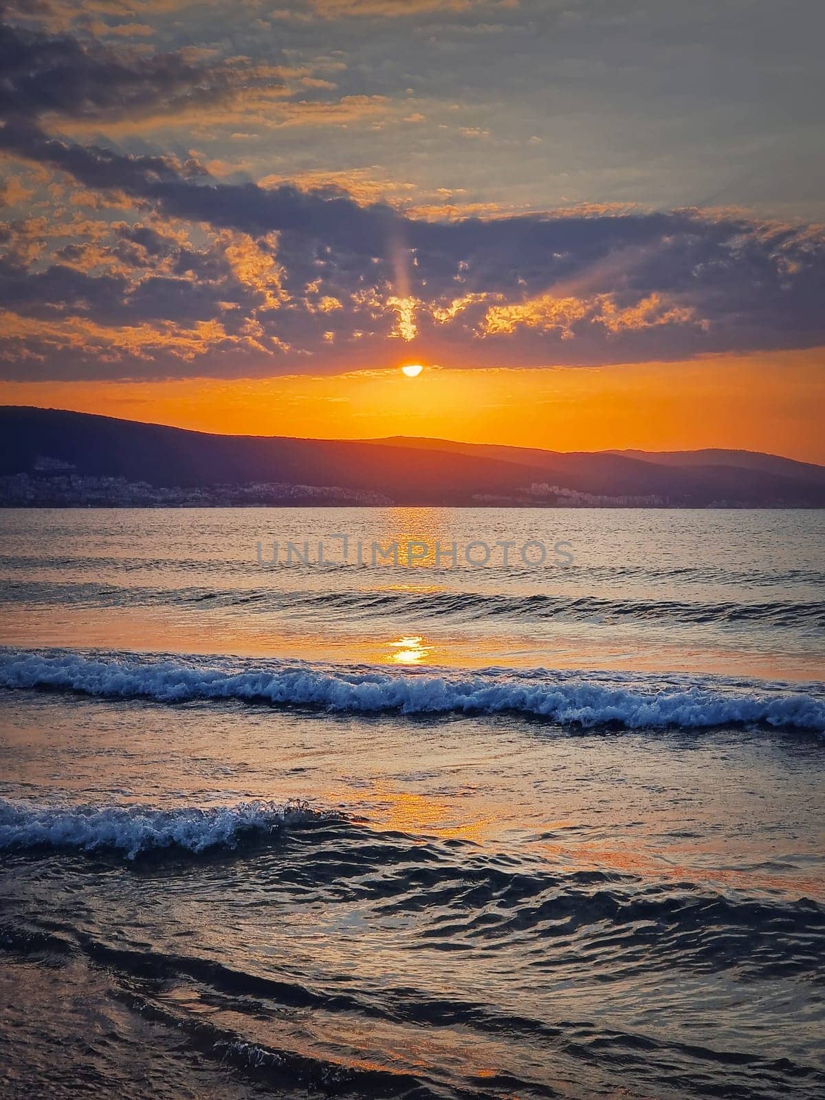 Vibrant sunrise at the Bulgarian coastline of Black Sea. Sunny beach resort, dawn scene with the sun rising up the hills on the horizon and foamy waves washing the sand shore