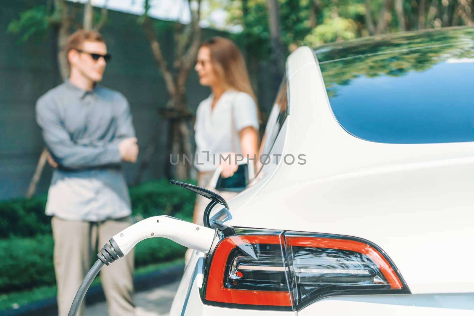 Young couple travel with EV electric car charging in green sustainable city outdoor garden in summer shows urban sustainability lifestyle by green clean rechargeable energy of electric vehicle innards