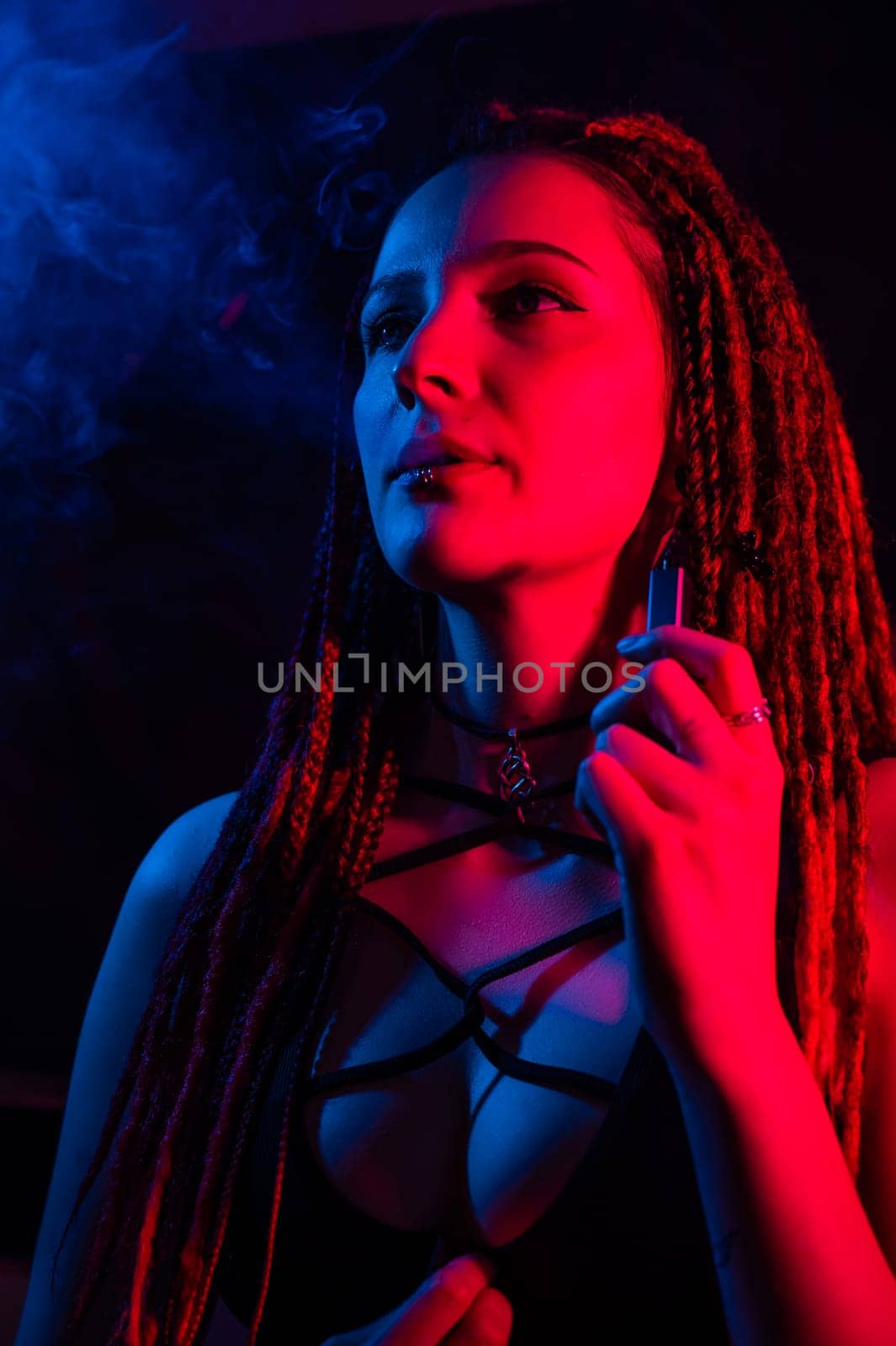 Caucasian girl with dreadlocks smokes a vape in red blue light