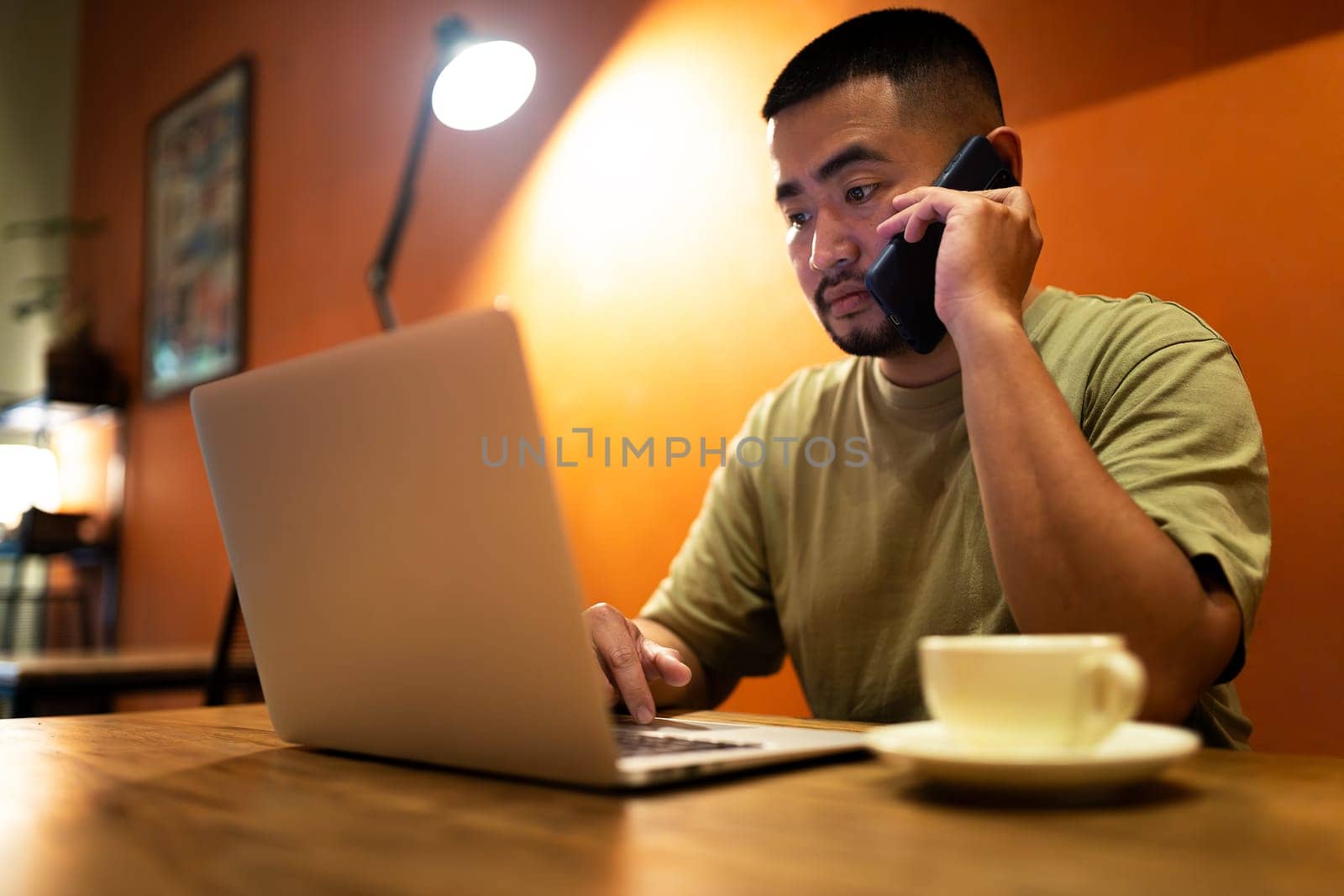 Asian man talking on the phone with client while working in coffee shop using laptop. by Hoverstock