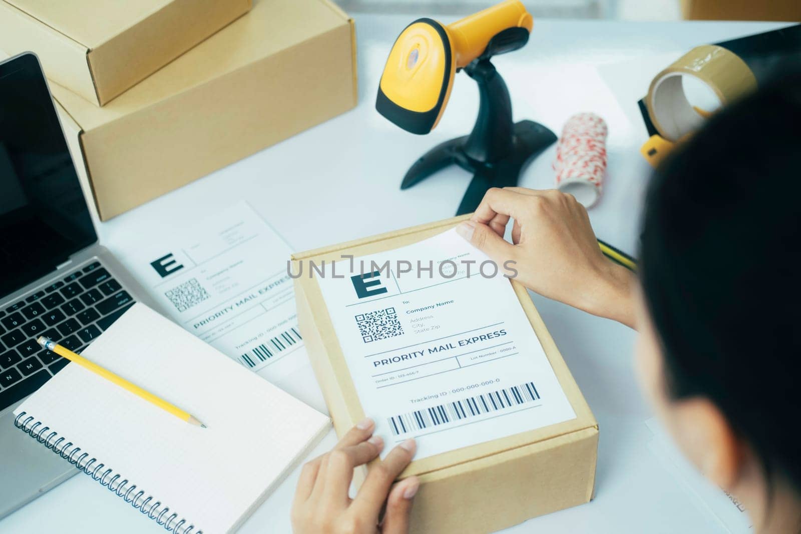 Close up of young female online business owner sitting at desk with laptop and boxes and putting shipping label on cardboard boxes or parcel of customer's order preparing for shipment and delivery.
