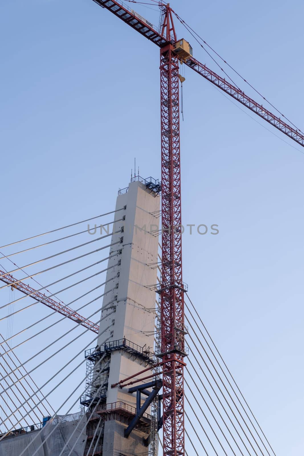 Crane and bridge construction against a blue sky background. Builders work on large construction sites, and there are many cranes working. There are a lot of cables on the bridge