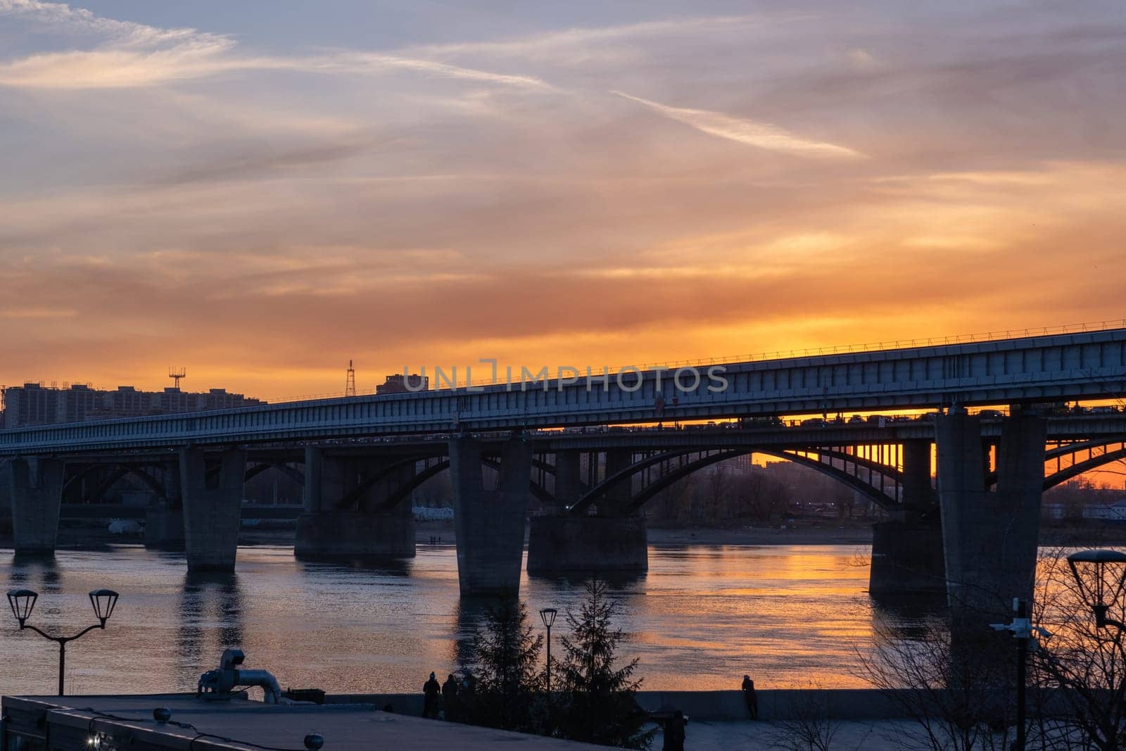 Beautiful view of the bridge over which cars drive at sunset. A river flows under the bridge, reflecting the sunset rays. Bridge in the city of Novosibirsk, Russia