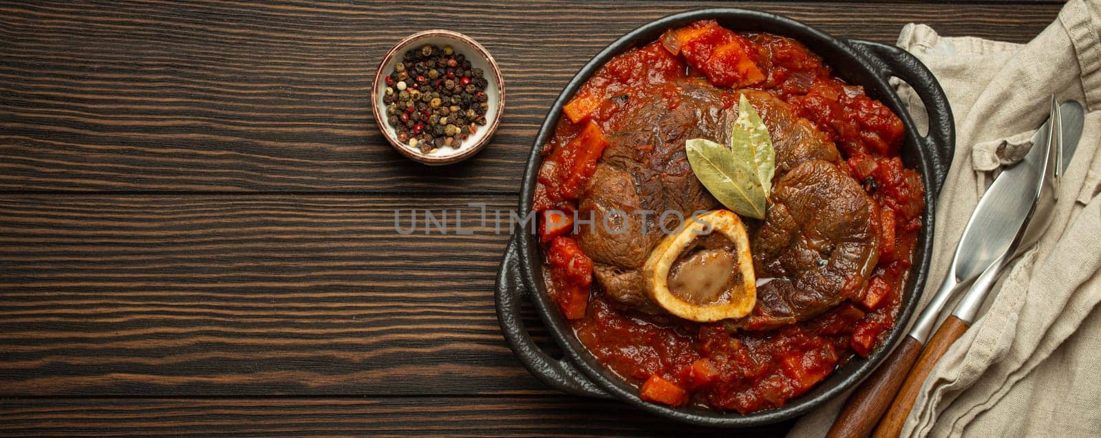 Traditional Italian dish Ossobuco all Milanese made with cut veal shank meat with vegetable tomato sauce served in black casserole pan top view on rustic brown wooden background, copy space by its_al_dente