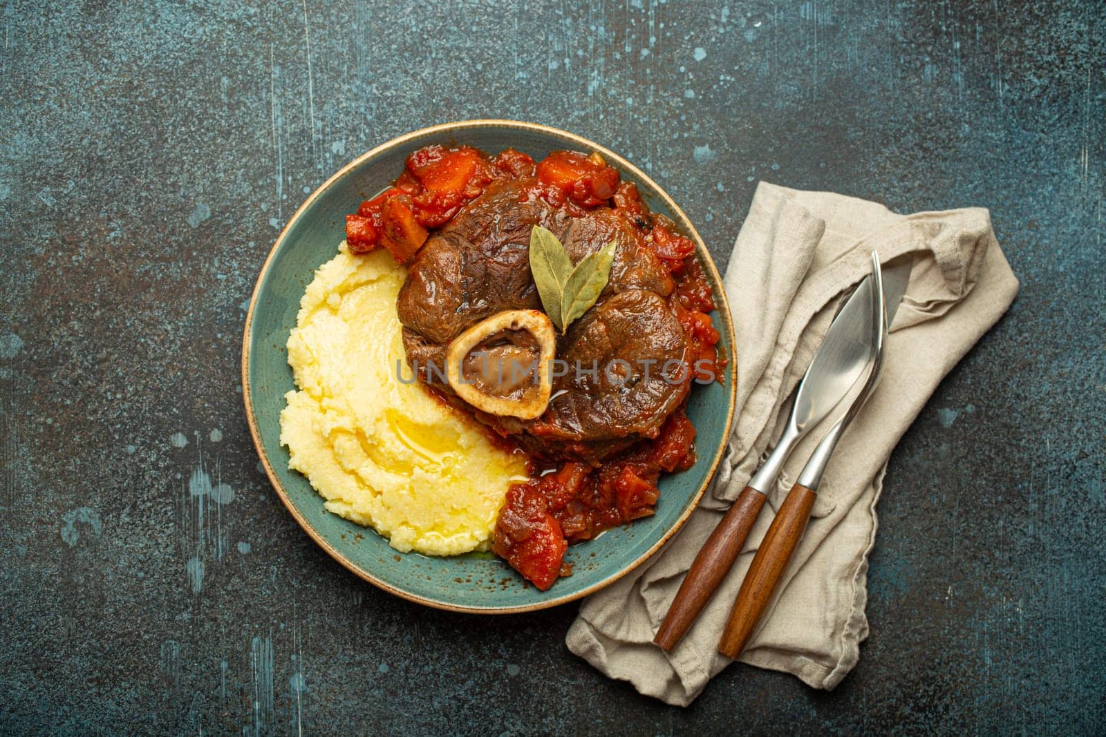 Traditional Italian dish Ossobuco all Milanese made with cut veal shank meat with vegetable tomato sauce served with corn polenta on ceramic plate top view on rustic stone background.