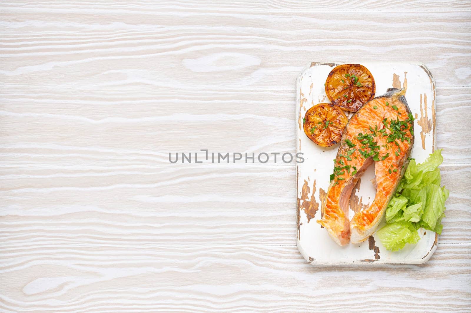 Grilled fish salmon steak and green salad with lemon served on white cutting board rustic wooden background top view, balanced diet or healthy nutrition meal with salmon and veggies, copy space.