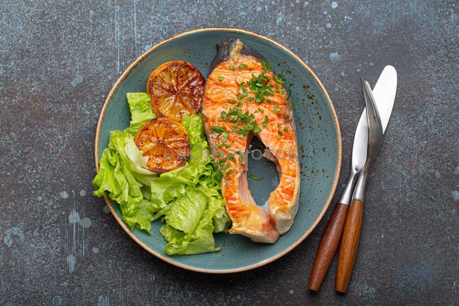 Grilled fish salmon steak and green salad with lemon on ceramic plate on rustic blue stone background top view, balanced diet or healthy nutrition meal with salmon and veggies.