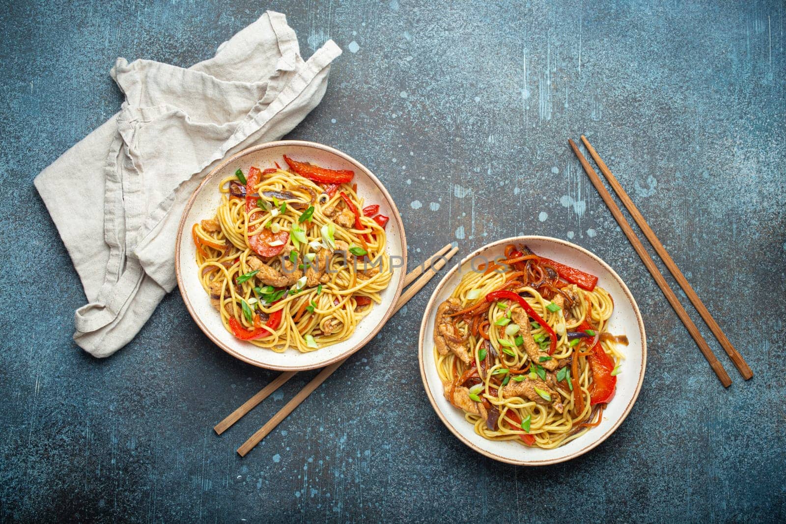 Two bowls with Chow Mein or Lo Mein, traditional Chinese stir fry noodles with meat and vegetables, served with chopsticks top view on rustic blue concrete background.