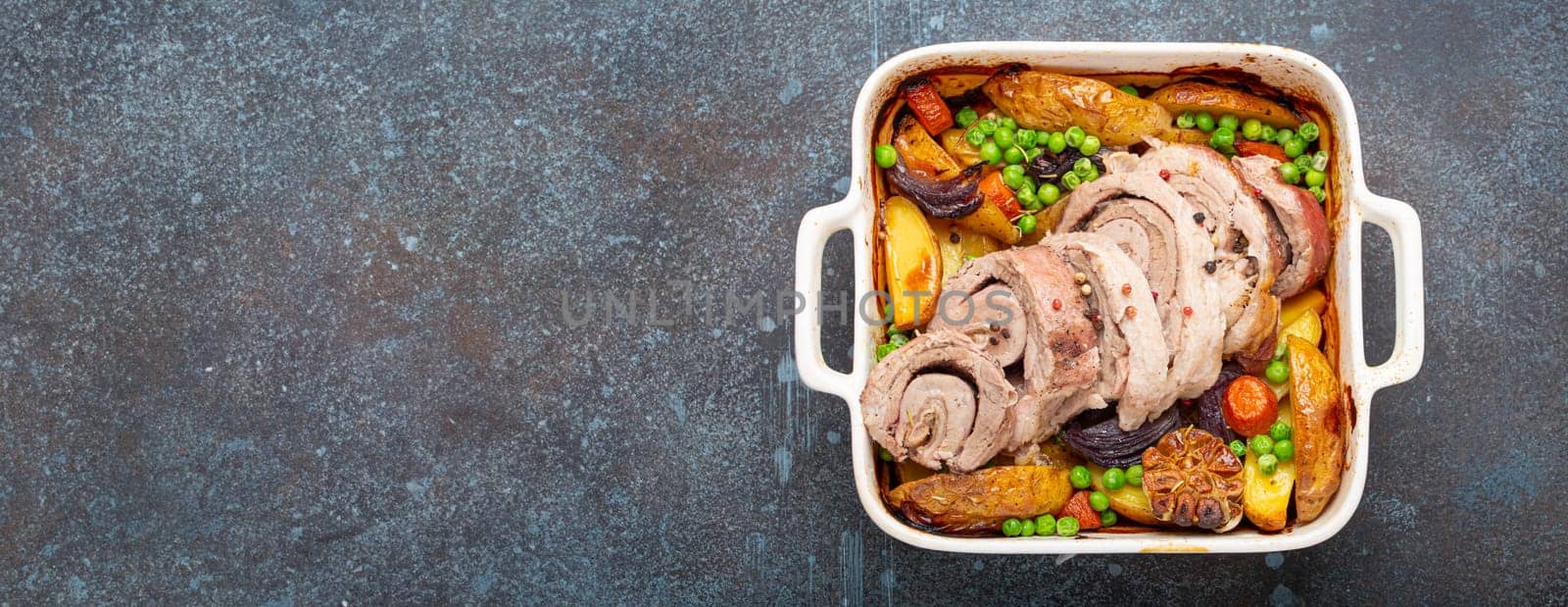 Rolled sliced pork roasted in white casserole dish with potatoes, vegetables and herbs on blue dark concrete rustic background top view. Baked pork roll with vegetables for dinner.