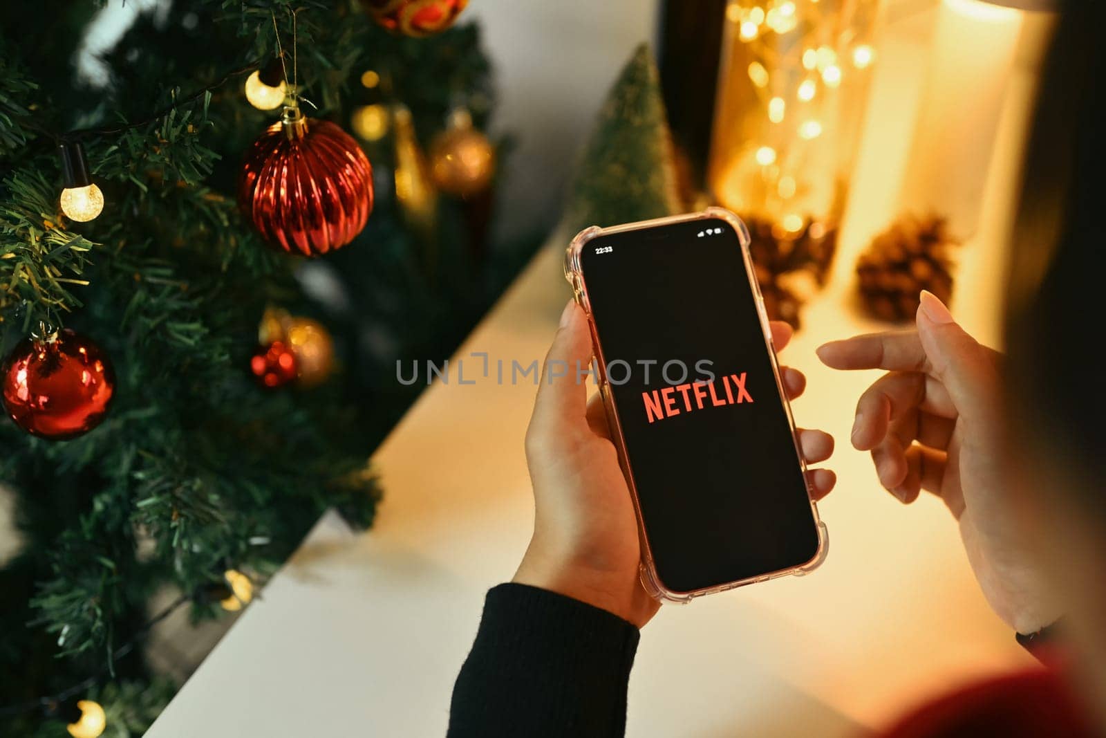 Chiang Mai, Thailand - Oct 31,2023: Woman holds a smartphone with the Netflix logo displayed on the screen near Christmas tree by prathanchorruangsak