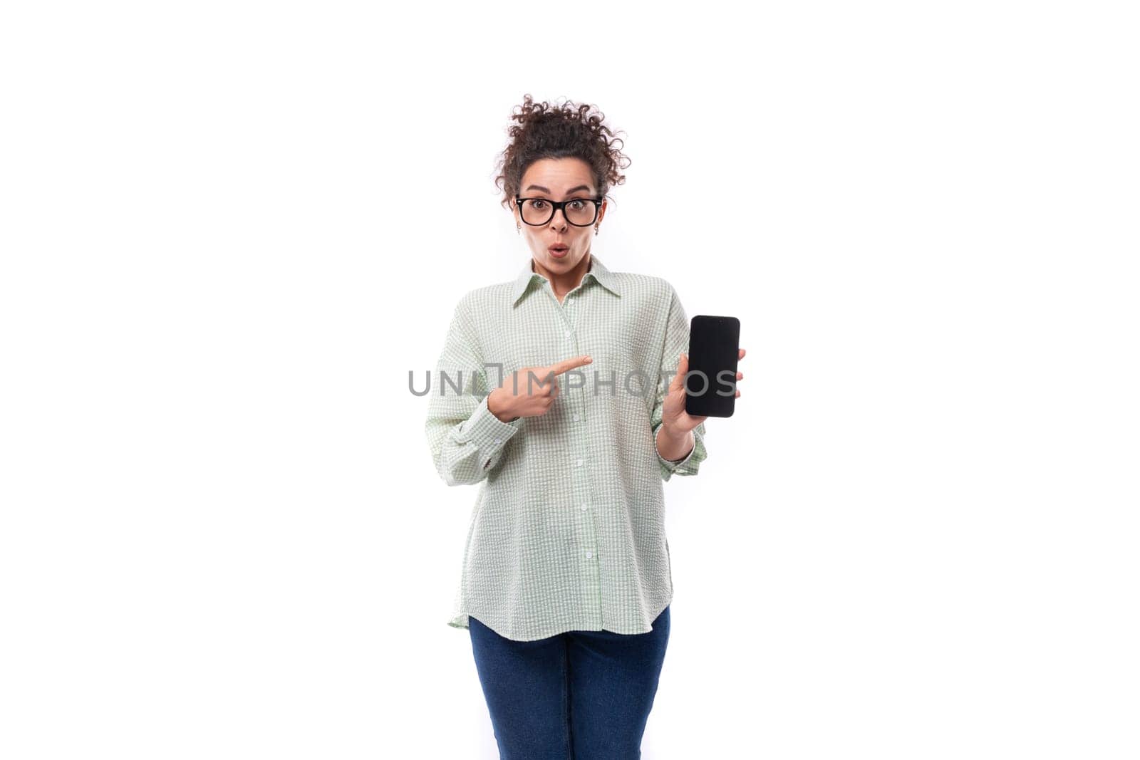 young successful teacher woman with curly black hair in a shirt and jeans shows the screen of a mobile phone in surprise.