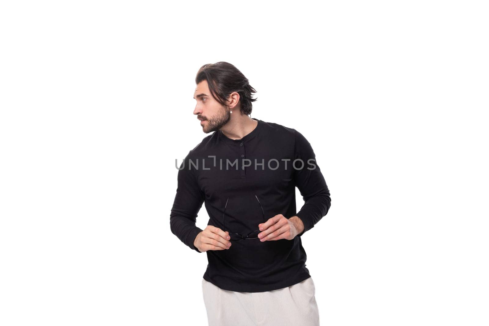 brutal young brunette man with a beard in a black turtleneck holds glasses for vision correction in his hands by TRMK