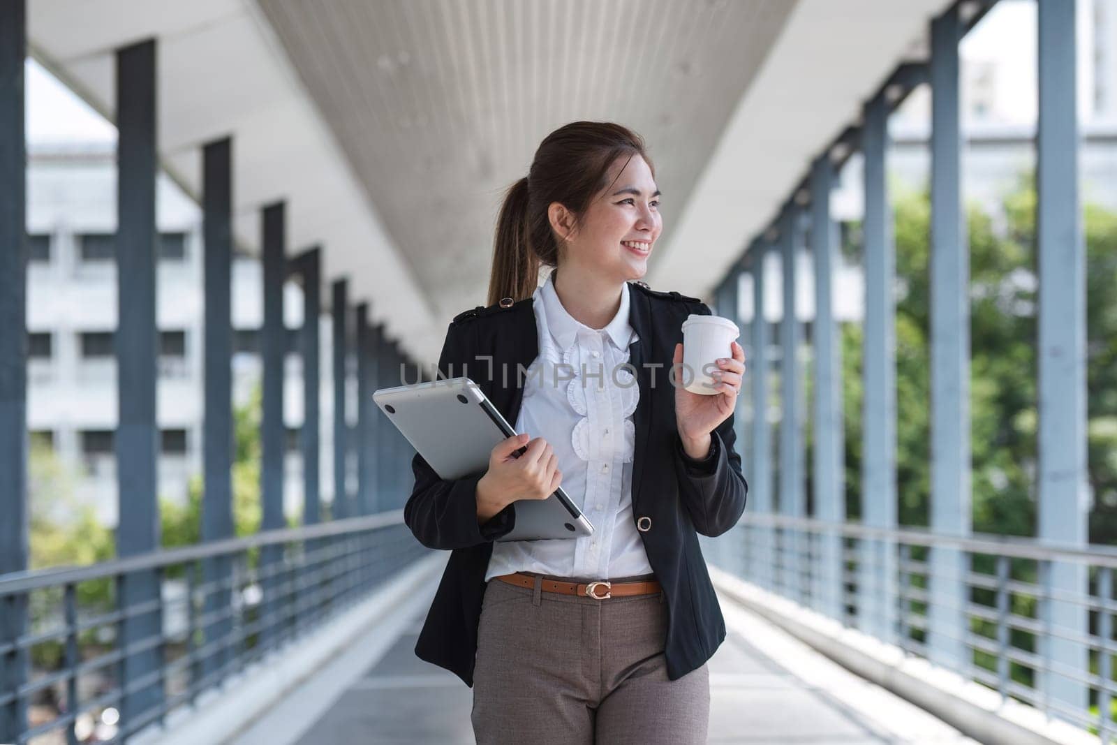 Asian businesswoman holding a laptop and a coffee mug. Smiling at the camera. Confident female employee holding a laptop standing in front of the camera on the background of a city skywalk. by wichayada