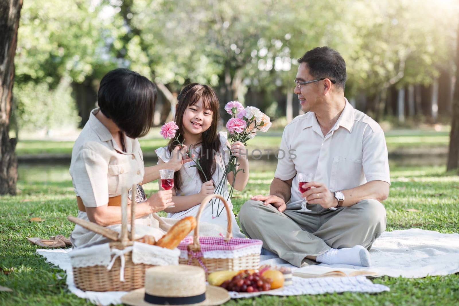 Family of senior couple and daughter picnicking in the park showing love Or reconnect after retirement in a relaxing park. An elderly man and a woman have fun on a mat in the backyard. by wichayada