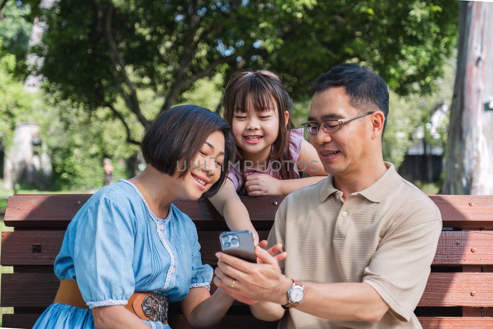 Kind and happy Asian senior grandparents enjoying looking at photos on their mobile phone in the park with their cute little granddaughter. On a clear day together lovely family by wichayada